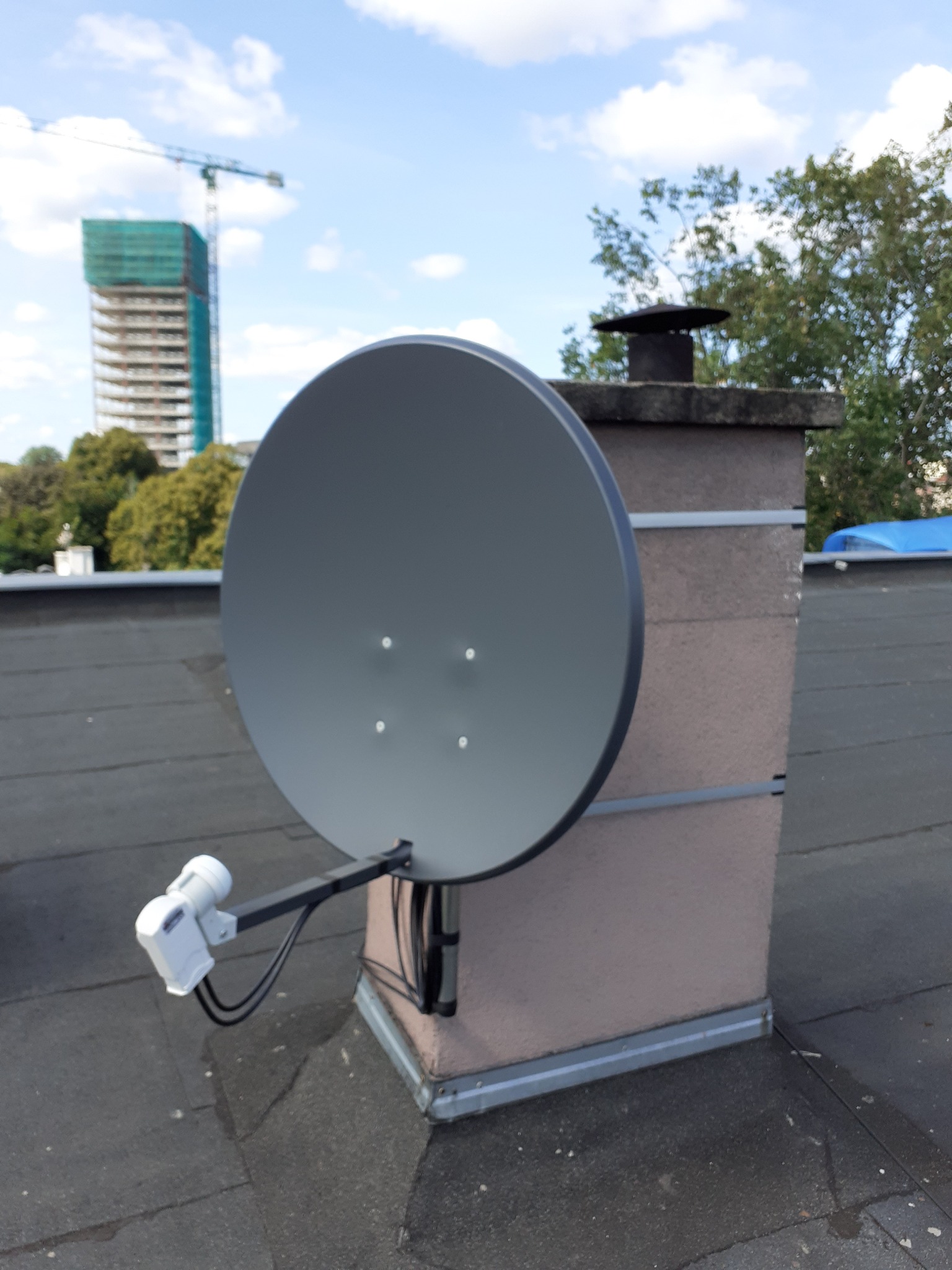 Szara antena satelitarna zamontowana na kominie na dachu pokrytym papą, w tle budynek w budowie i błękitne niebo z chmurami.