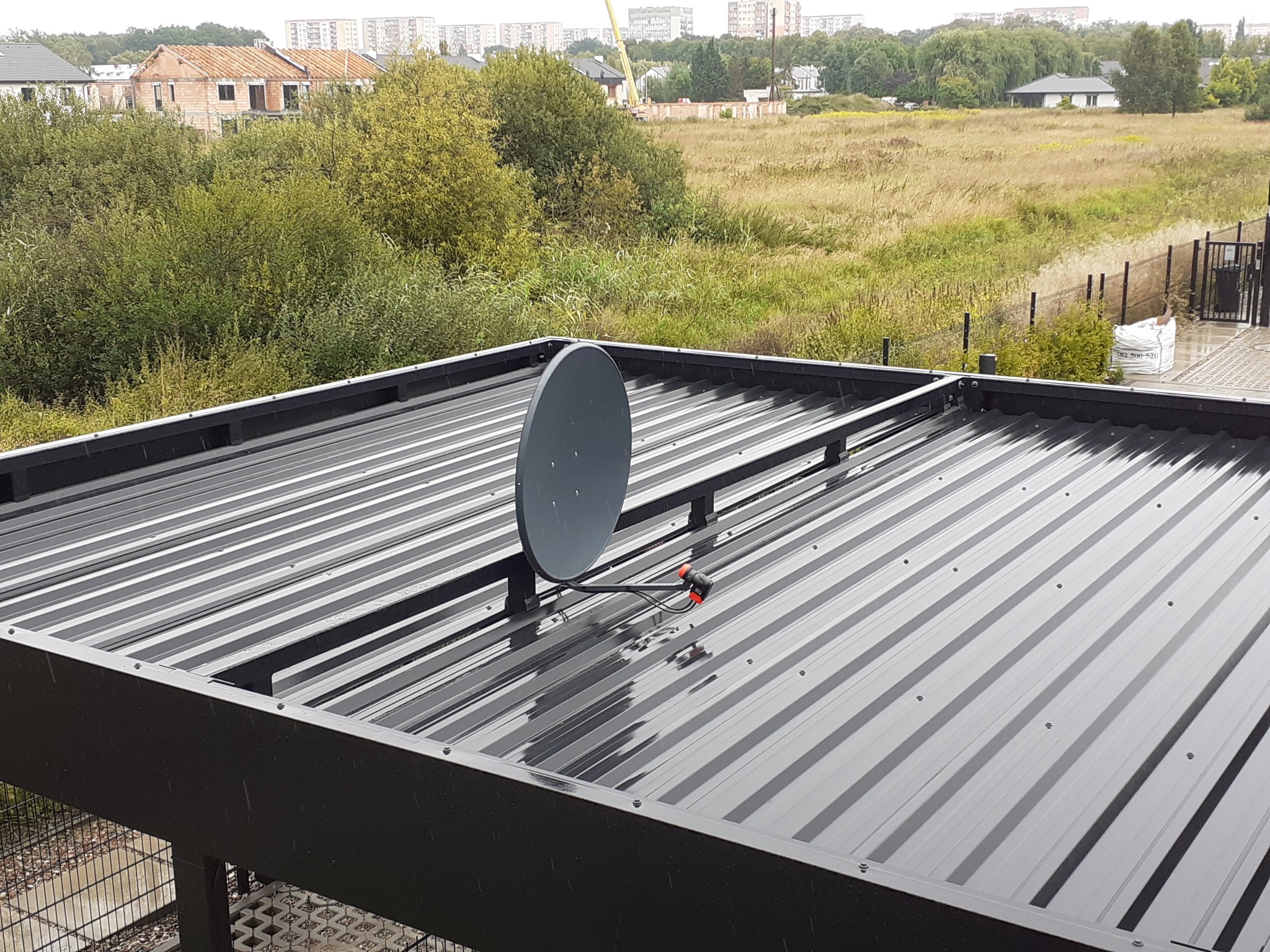 Szara antena satelitarna zamontowana na lśniącej, metalowej konstrukcji dachu z widocznymi budynkami mieszkalnymi i zielenią w tle.
