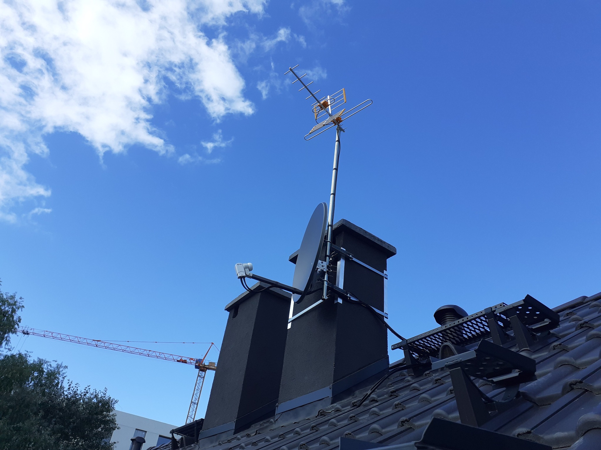 Antena telewizyjna i satelitarna zamontowana na kominie na tle błękitnego nieba z chmurami. W tle widoczny żuraw budowlany.