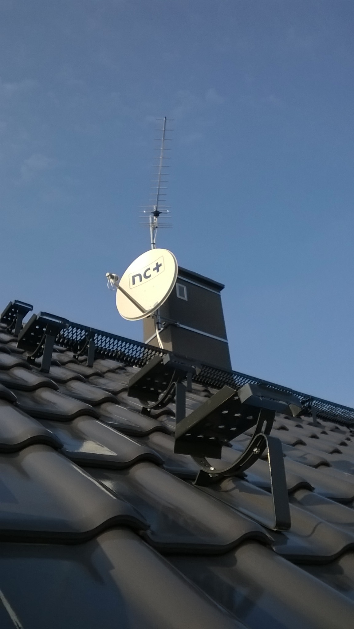 Antena satelitarna nc+ i antena naziemna zamontowane na dachu z dachówki ceramicznej, widok z dołu na tle błękitnego nieba.