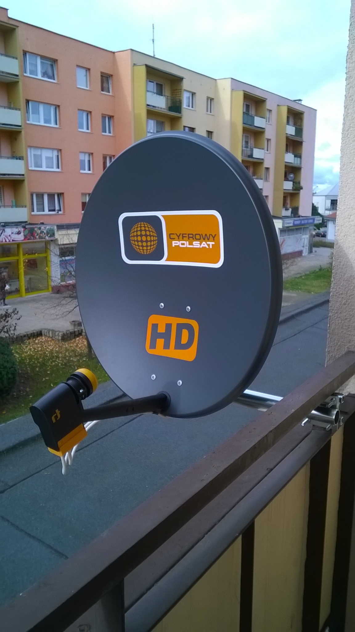 Szara antena satelitarna Cyfrowy Polsat HD zamontowana na balkonie z widokiem na blok mieszkalny w tle.