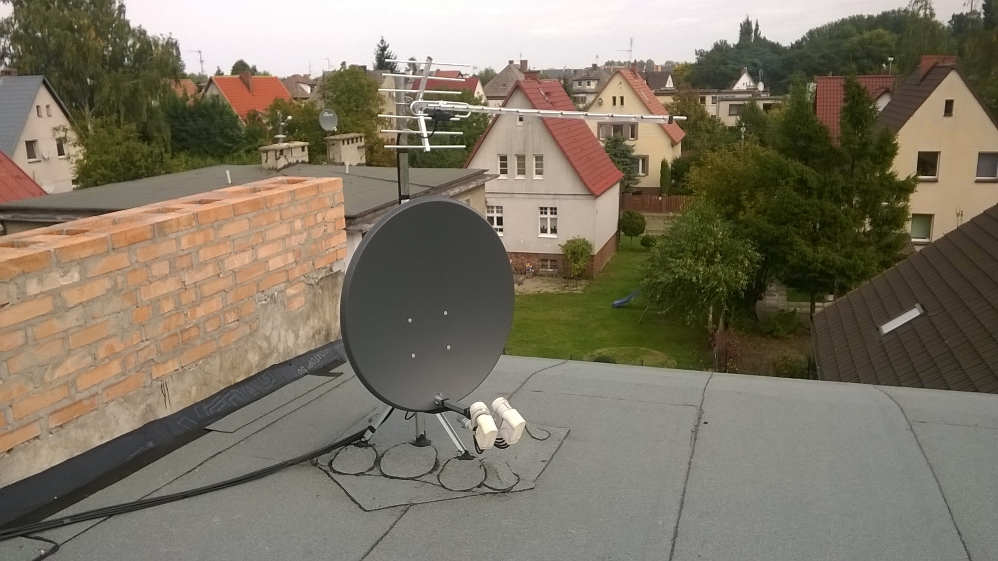 Antena satelitarna i antena kierunkowa UHF zamontowane na płaskim dachu budynku, widok na okoliczne domy i zieleń.