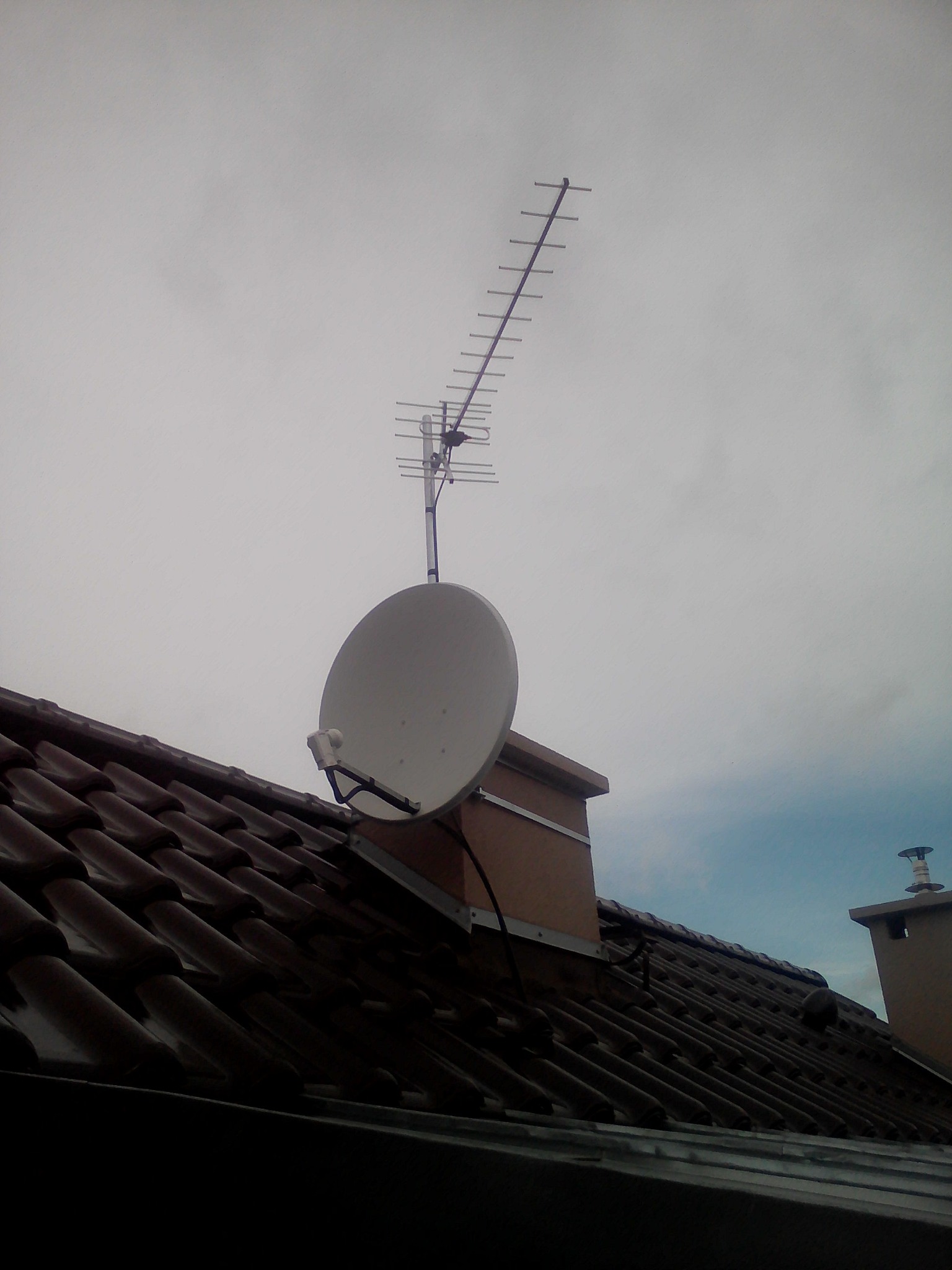 Antena satelitarna i antena naziemna zamontowane na kominie na brązowym dachu pokrytym dachówką ceramiczną, widok z dołu na pochmurne niebo.
