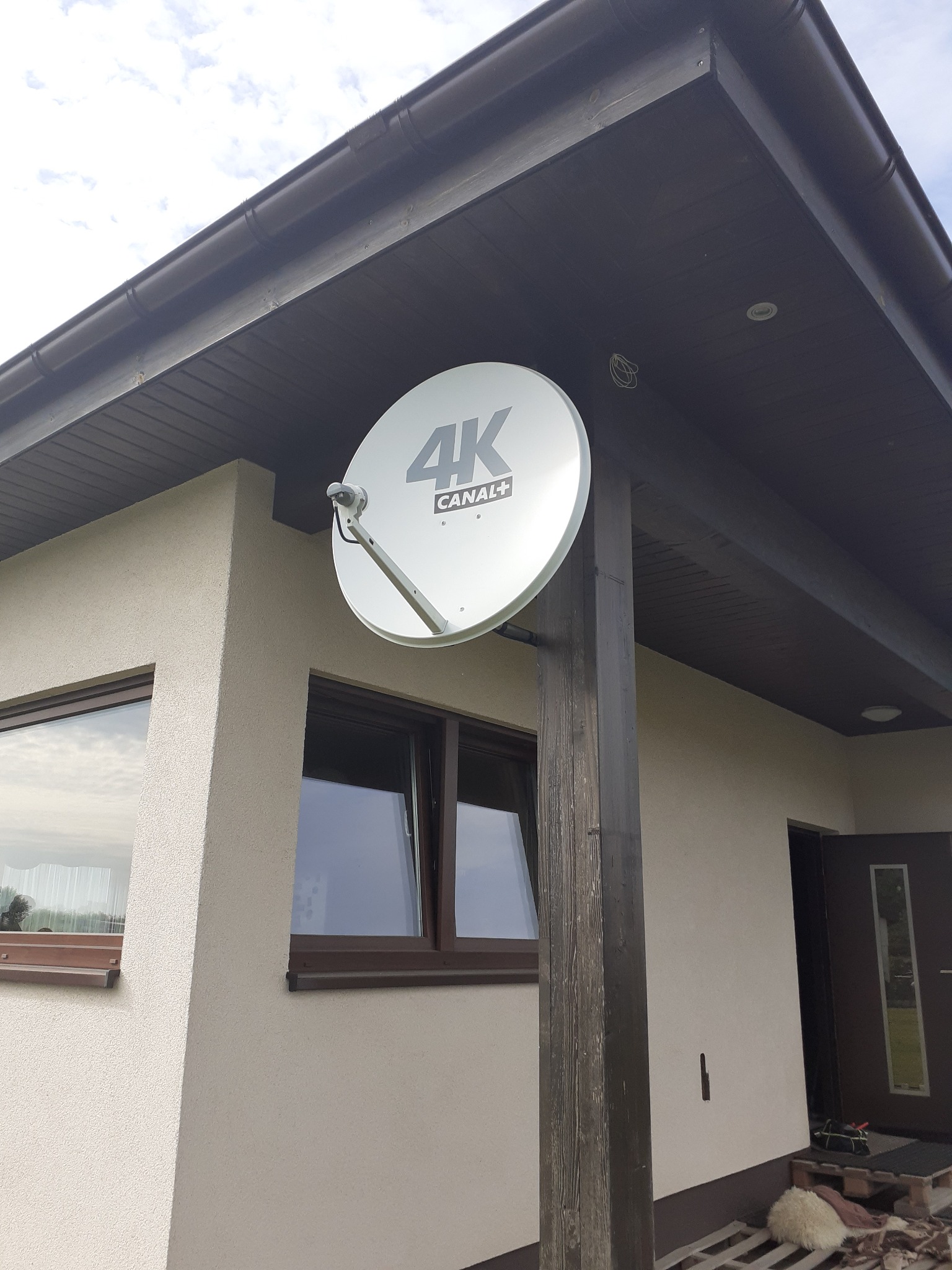 Biała antena satelitarna 4K Canal+ zamontowana na drewnianym słupie przy elewacji budynku z brązowymi oknami i podbitką dachu.