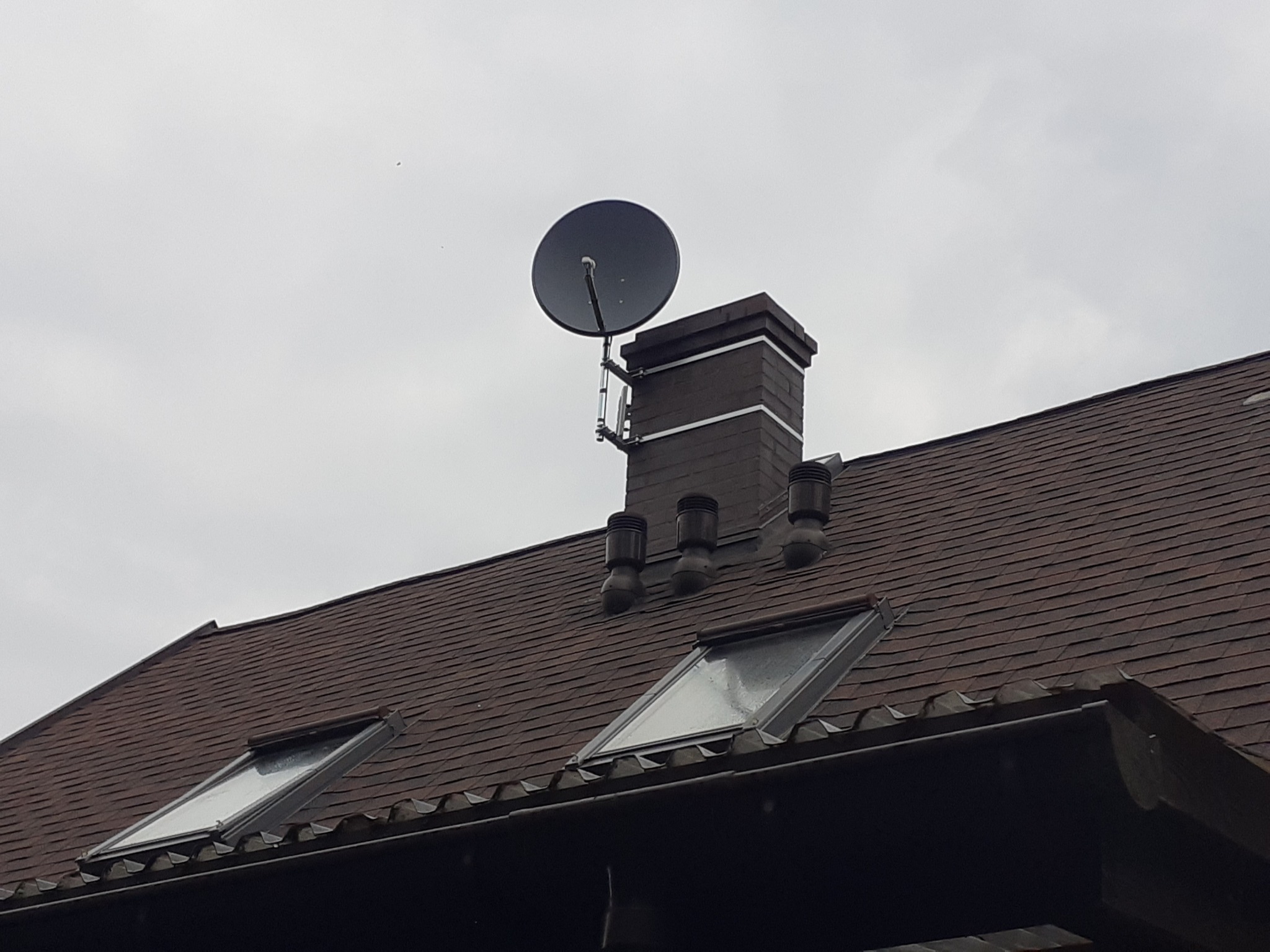 Antena satelitarna zamontowana na kominie z cegły na brązowym dachu z oknami dachowymi.
