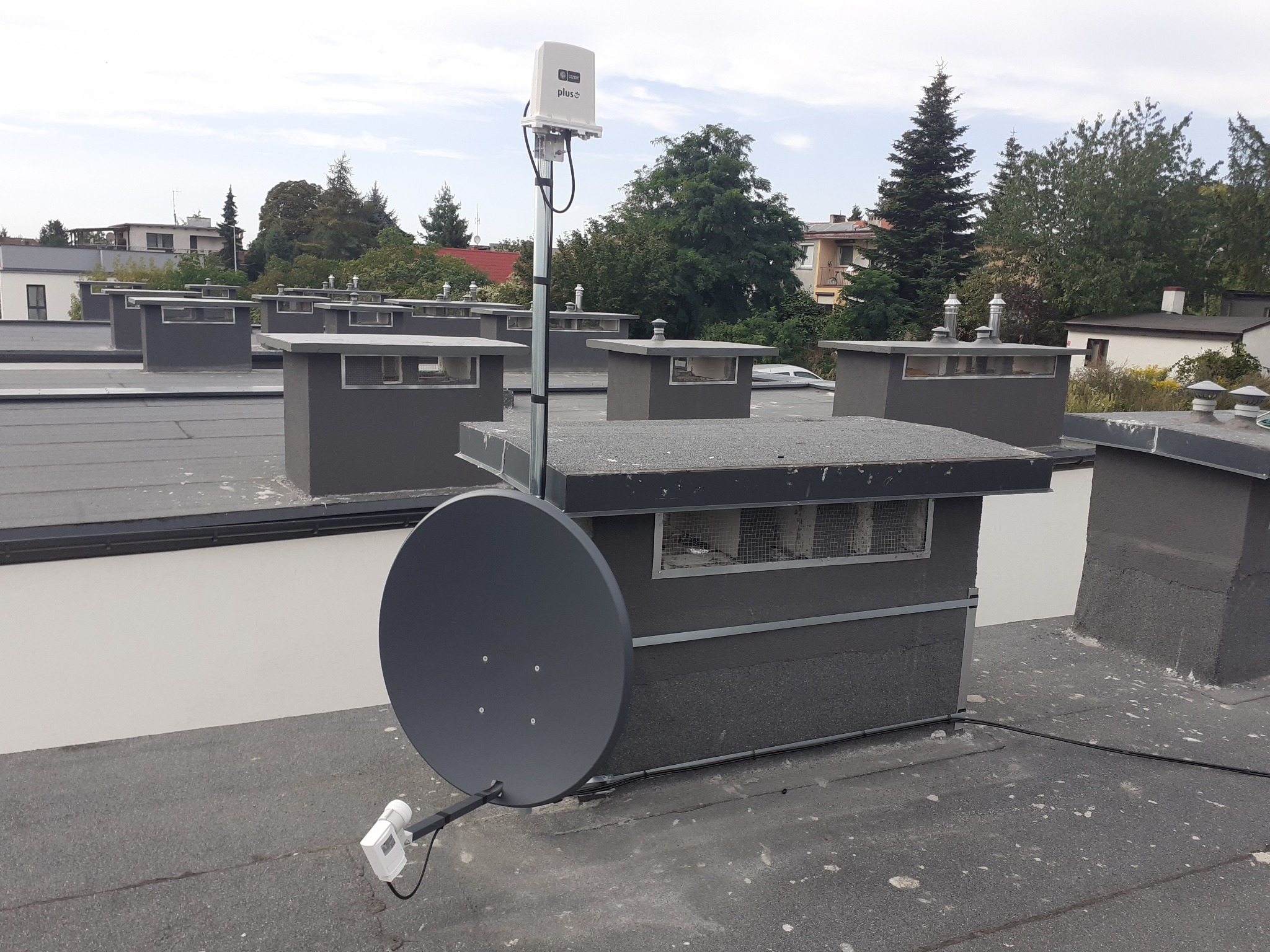Antena satelitarna i antena operatora Plus zamontowane na dachu budynku z kominami wentylacyjnymi w tle.