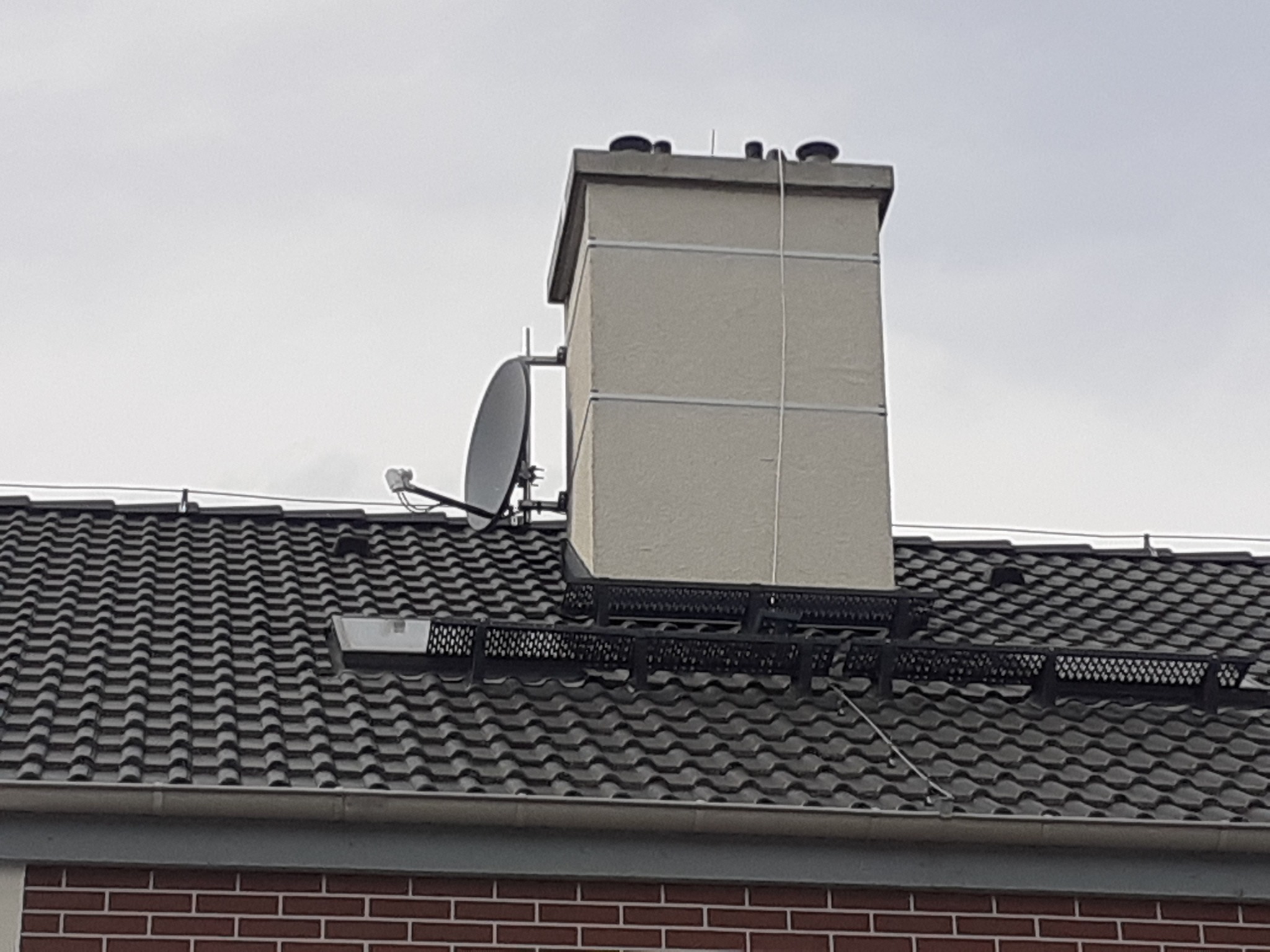 Antena satelitarna zamontowana na dachu obok komina z szarymi dachówkami, widok z dołu na tle pochmurnego nieba.