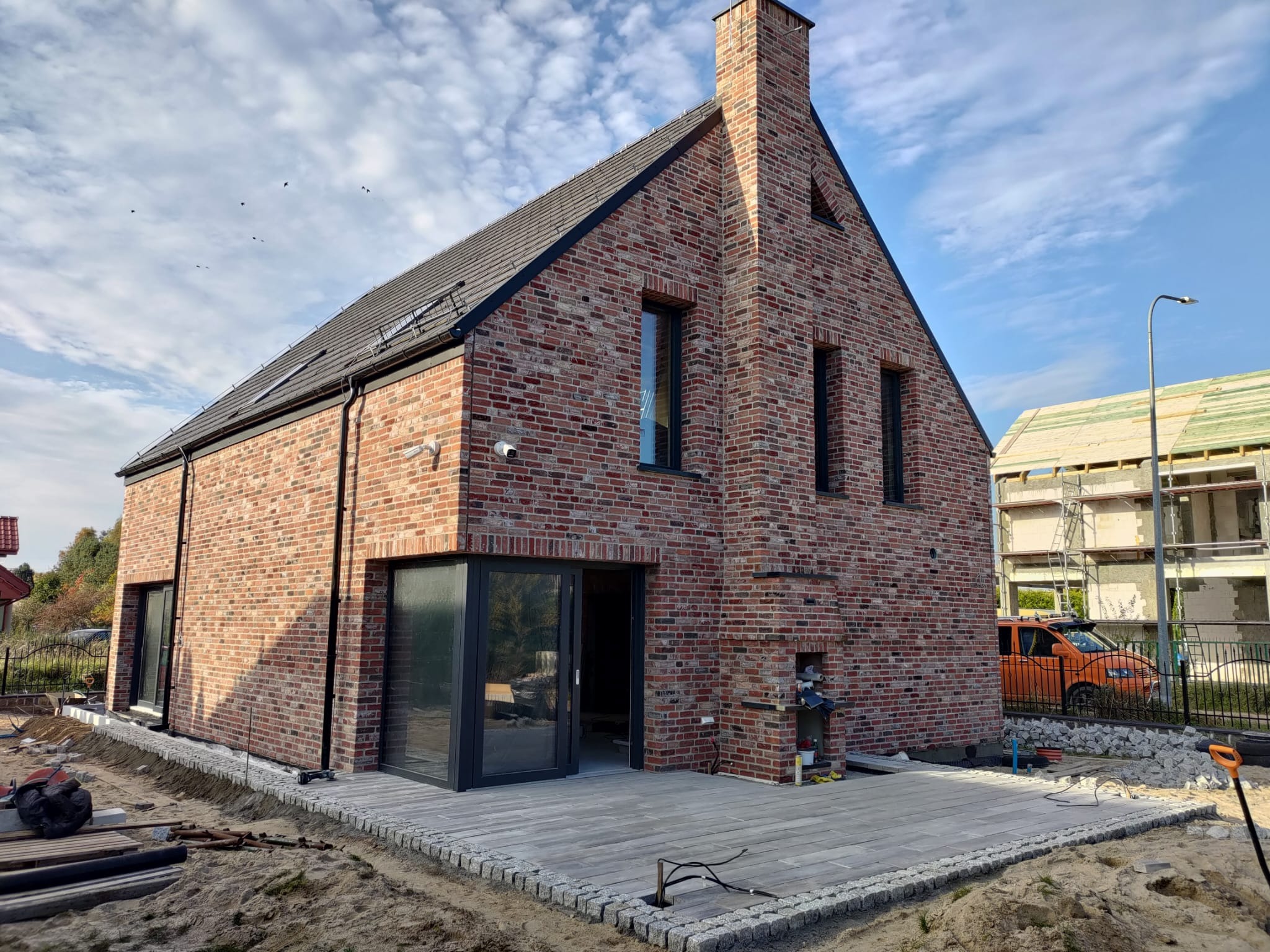 Nowoczesny dom z czerwonej cegły z ciemną stolarką okienną i brukowanym tarasem. Widoczne detale wykończenia elewacji i otoczenia domu.