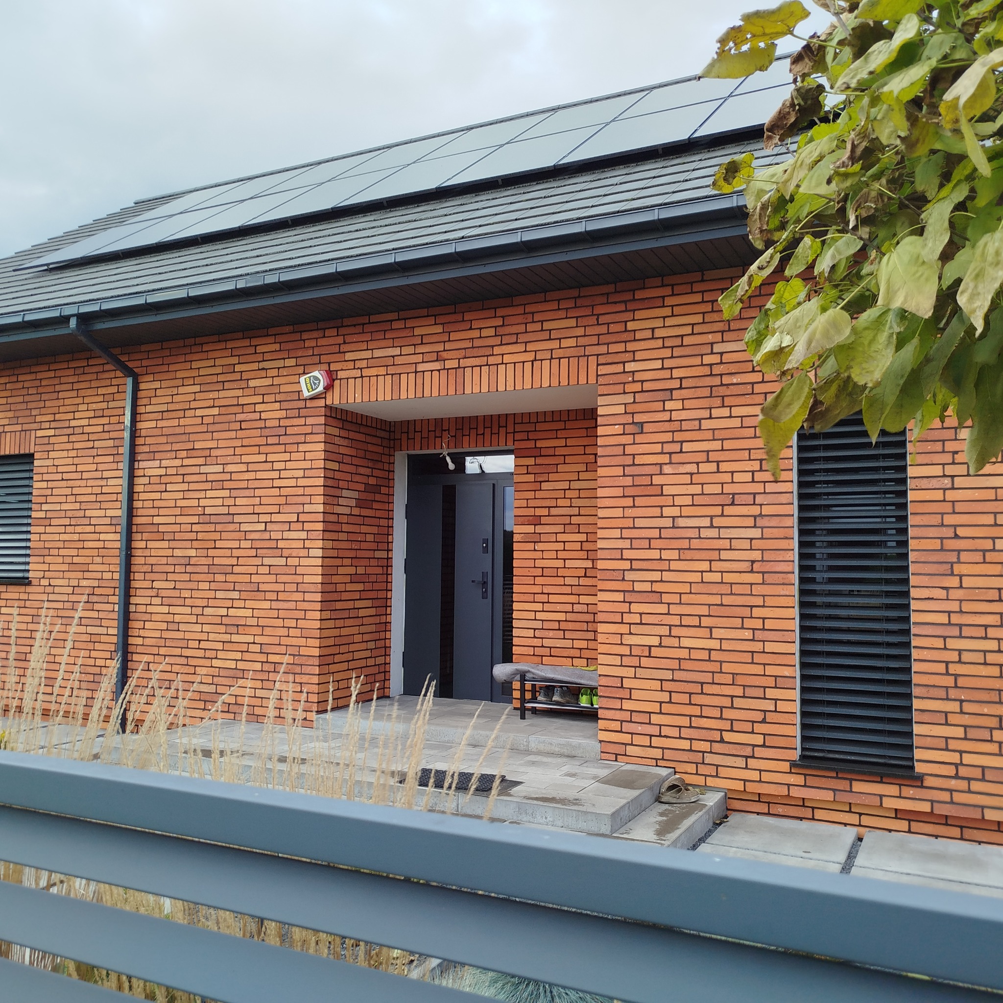 Nowoczesny dom z ceglaną elewacją, panelami słonecznymi na dachu i szarą stolarką okienną. Widok zza ogrodzenia, z elementami roślinności w kadrze.