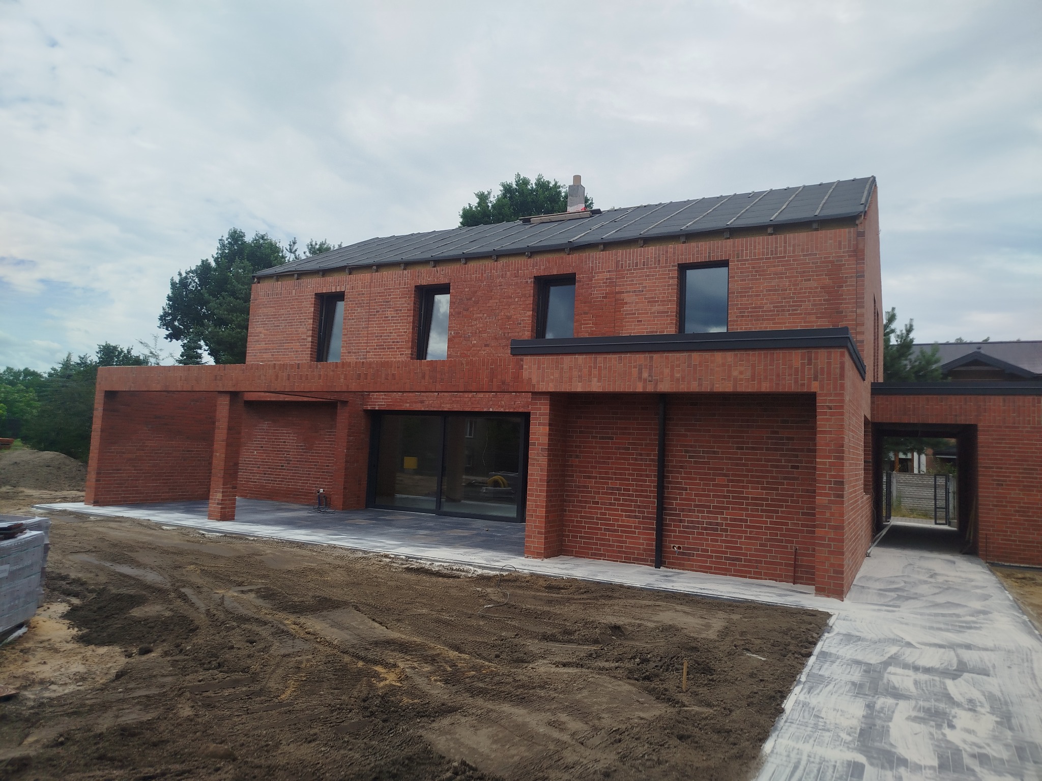 Nowoczesny dom z czerwonej cegły z dużymi oknami i płaskim dachem, otoczony ziemią. Widoczny fragment chodnika i zieleń w tle. Minimalistyczny design.