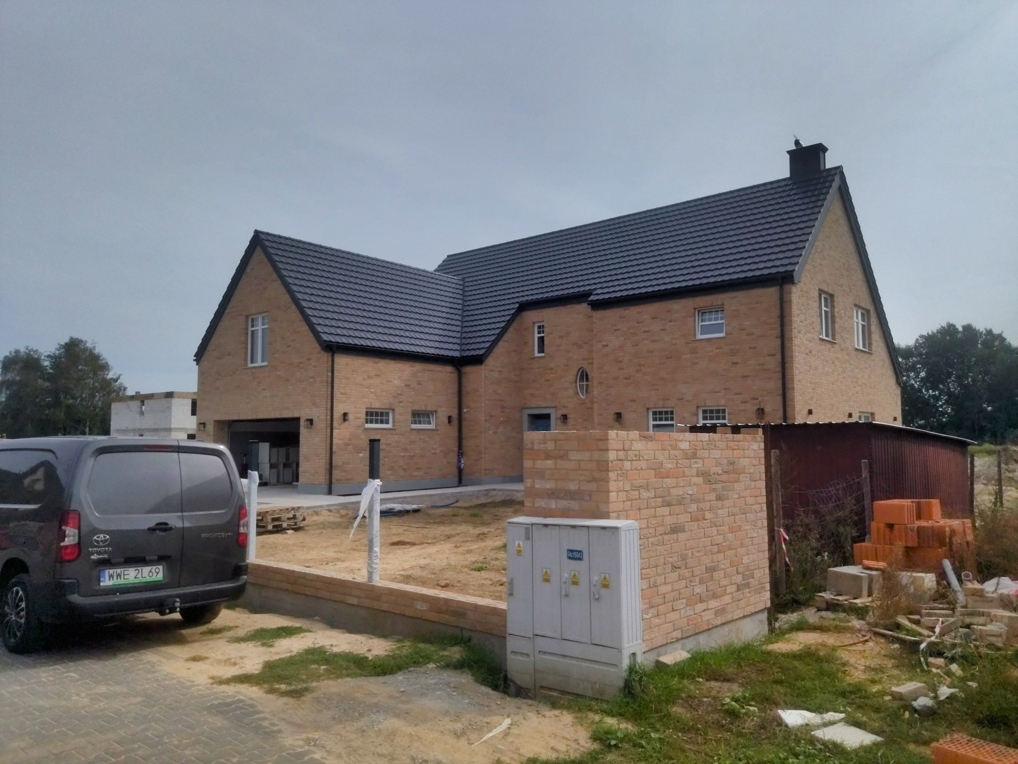 Nowoczesny dom z cegły klinkierowej z ciemną dachówką, widoczny z zewnątrz, z samochodem dostawczym zaparkowanym obok. Częściowo wykończony teren wokół budynku.