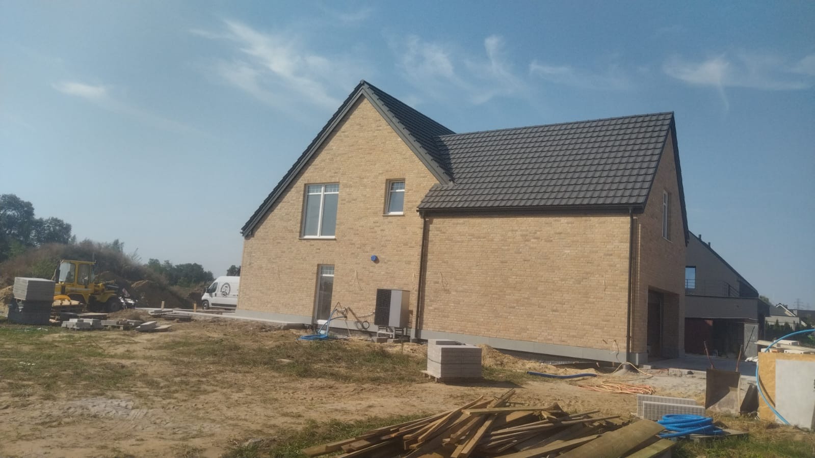 Nowoczesny dom z jasnej cegły klinkierowej, grafitowy dach, widoczne elementy placu budowy: palety z cegłami, rury, maszyna budowlana. Błękitne niebo w tle.