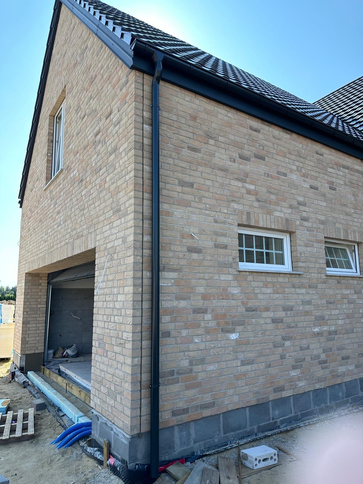 Nowoczesny dom w trakcie budowy z beżowej cegły, widoczne okna z białymi ramami, czarna rynna i fragment dachu. W tle błękitne niebo i elementy placu budowy.