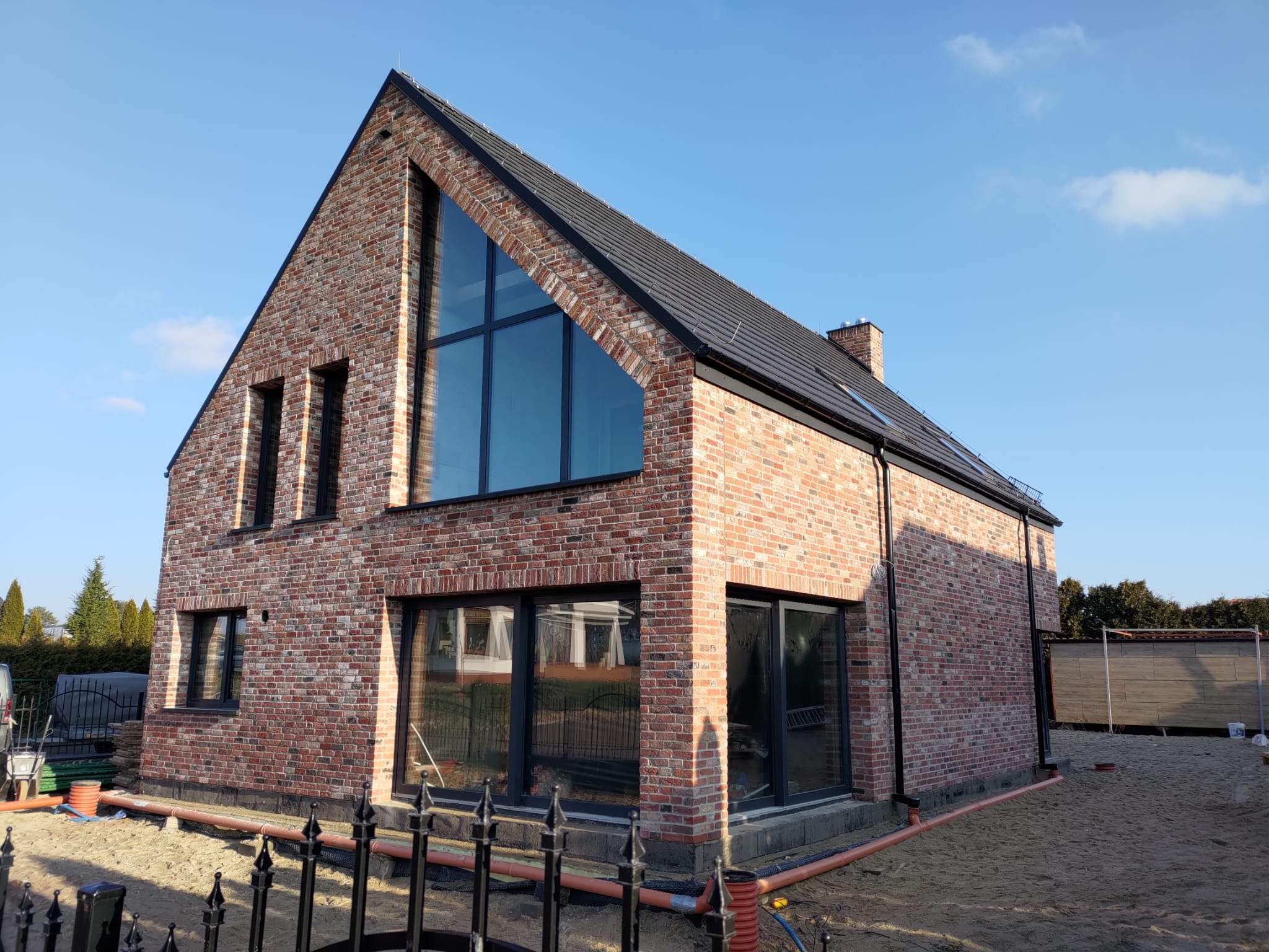 Dwukondygnacyjny dom z czerwonej cegły z dużym trójkątnym oknem na szczycie, czarną stolarką okienną i dachem z dachówki, otoczony piaskiem i czarnym ogrodzeniem.