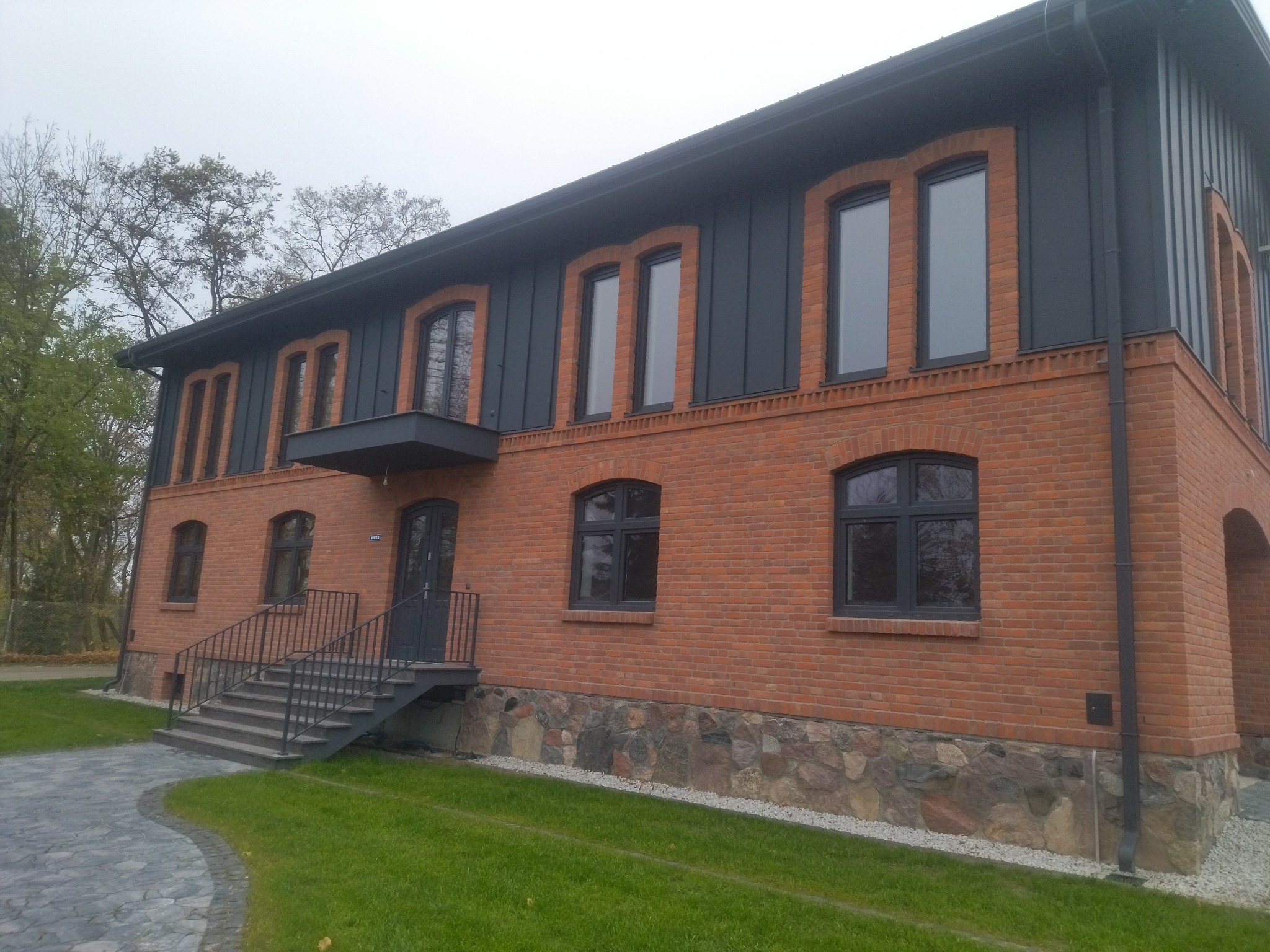 Dwukondygnacyjny budynek z cegły klinkierowej z ciemnoszarą elewacją i oknami w łukowych obramowaniach. Widoczne schody wejściowe i fragment trawnika.