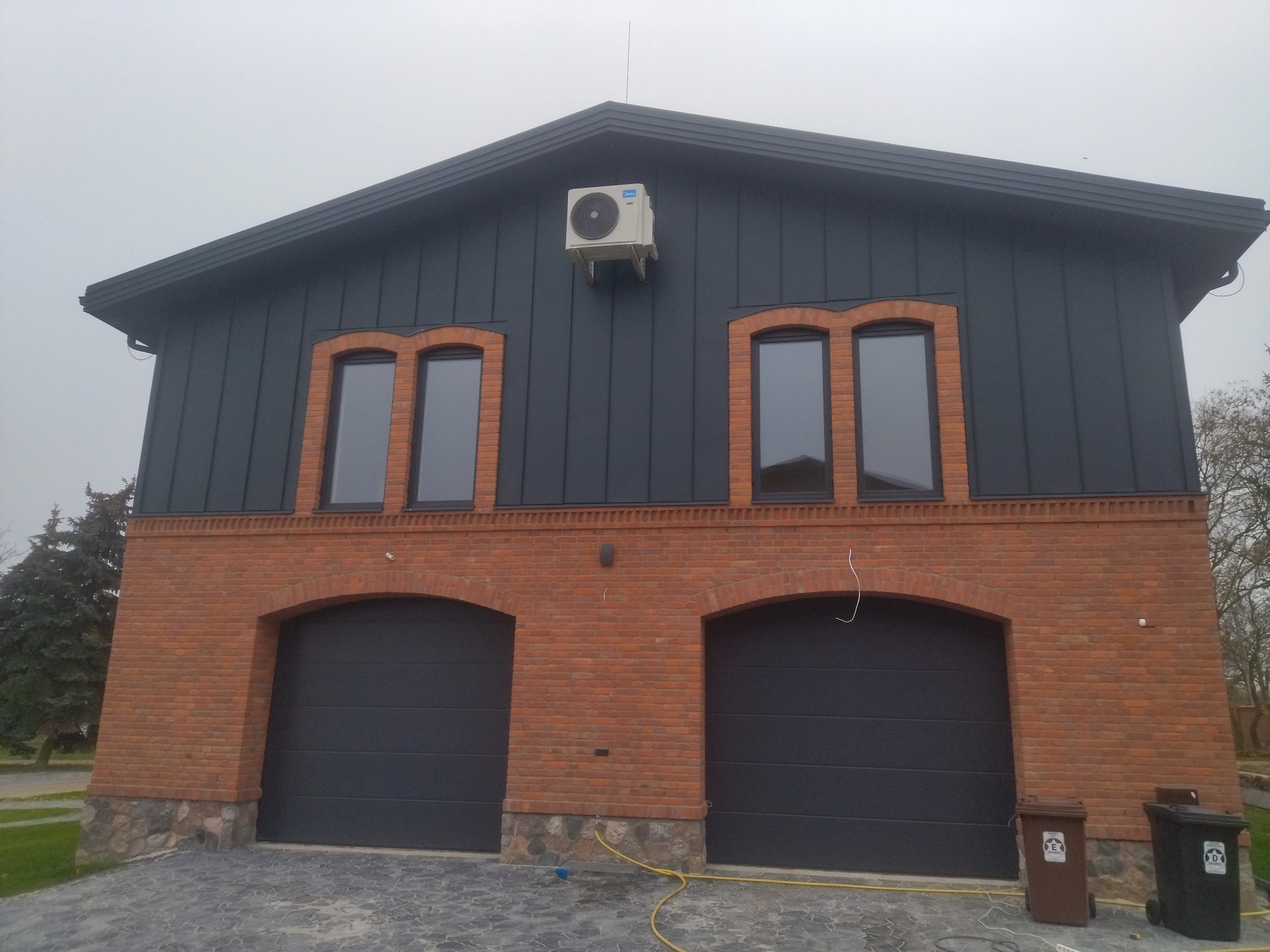 Dwukondygnacyjny budynek z ciemną elewacją i ceglanym parterem, z dwoma garażami i klimatyzatorem na ścianie. Widoczne okna z ceglanymi obramowaniami.