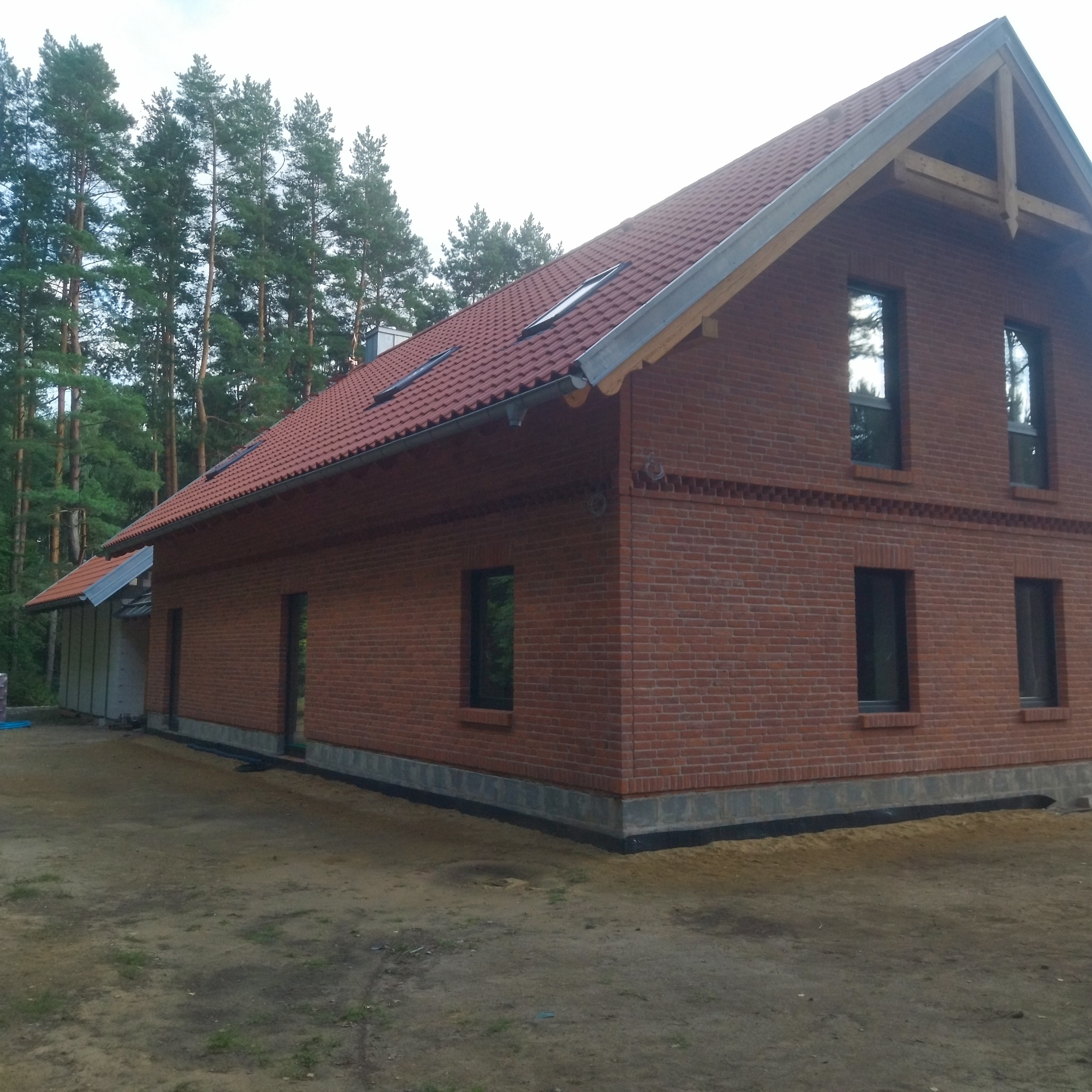Ceglany dom jednorodzinny z czerwoną dachówką, otoczony lasem. Widoczna elewacja z detalami z cegły klinkierowej i fragmentem ocieplonej ściany. Ujęcie z poziomu gruntu.