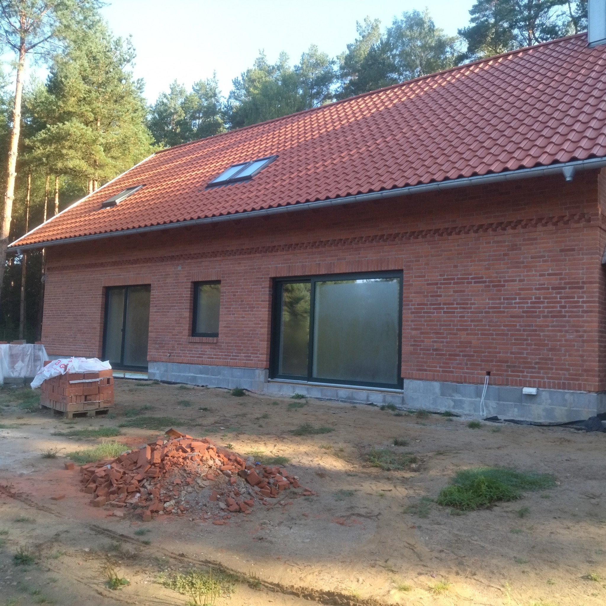 Nowy dom z cegły klinkierowej z czerwoną dachówką, widoczne okna dachowe i duże okna. Na pierwszym planie sterta cegieł i paleta z cegłami. W tle drzewa.