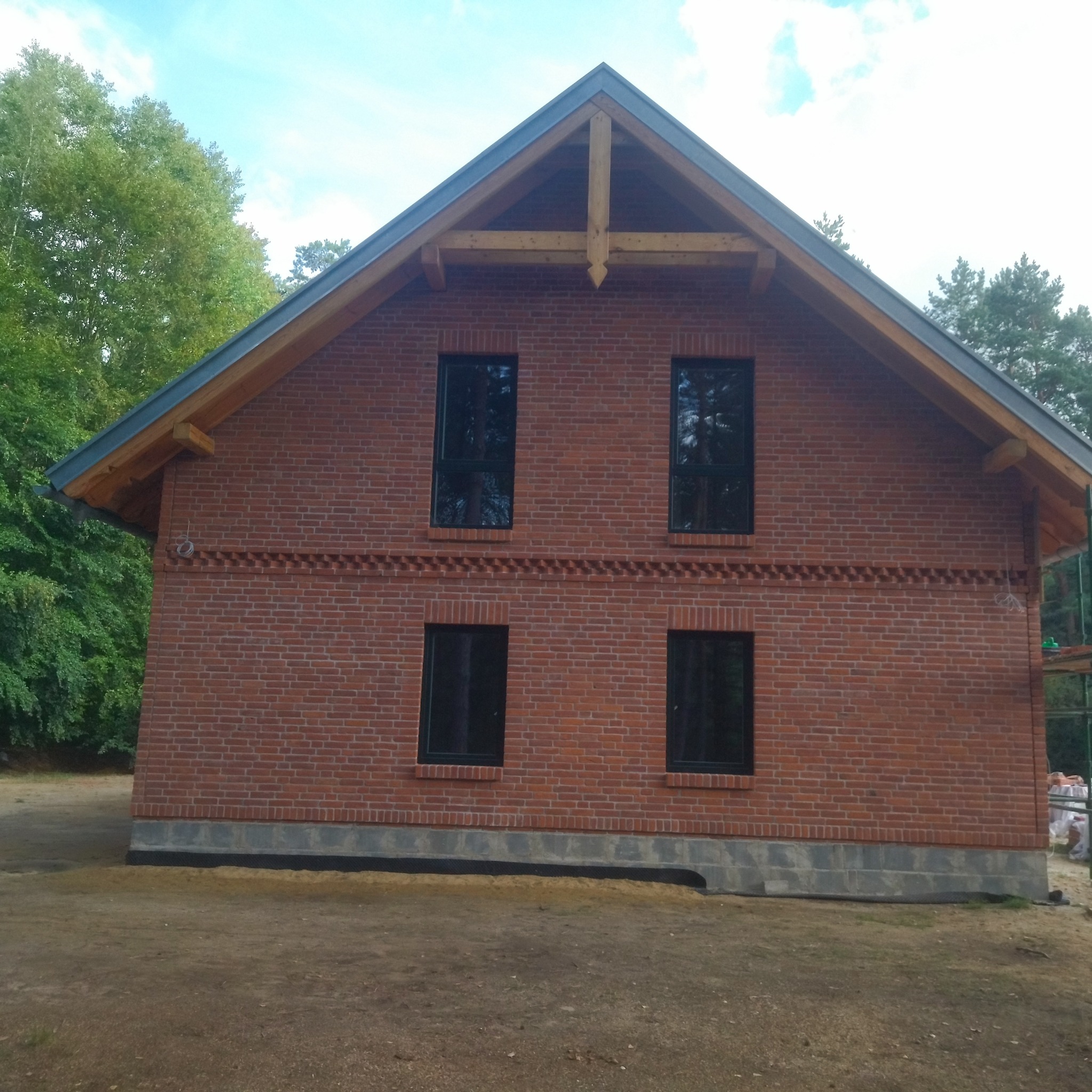 Dwukondygnacyjny dom z czerwonej cegły z czarnymi oknami i drewnianą konstrukcją dachu, otoczony zielenią. Widoczny fragment fundamentu i elewacji.