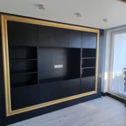 Czarna zabudowa meblowa z półkami i szafkami, obramowana złotą ramą, w nowoczesnym wnętrzu z widokiem na zewnątrz.