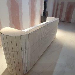 Fitino Jakub Małysek - Biała, zaokrąglona lada recepcyjna z pionowymi żłobieniami, w surowym stanie, w trakcie montażu w pomieszczeniu z przygotowanymi ścianami do malowania.