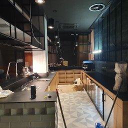 Fitino Jakub Małysek - Montaż nowoczesnej kuchni w lokalu gastronomicznym: widoczne niedokończone szafki, blaty robocze, instalacja oświetlenia, pracownik na drabinie i częściowo zakryta podłoga z wzorem geometrycznym.