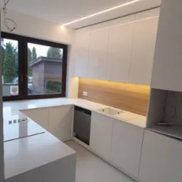 Nowoczesna, biała kuchnia z drewnianym panelem nad blatem, oświetleniem LED podszafkowym i dużym oknem z widokiem na zieleń, z niedokończonymi pracami elektrycznymi widocznymi na suficie.