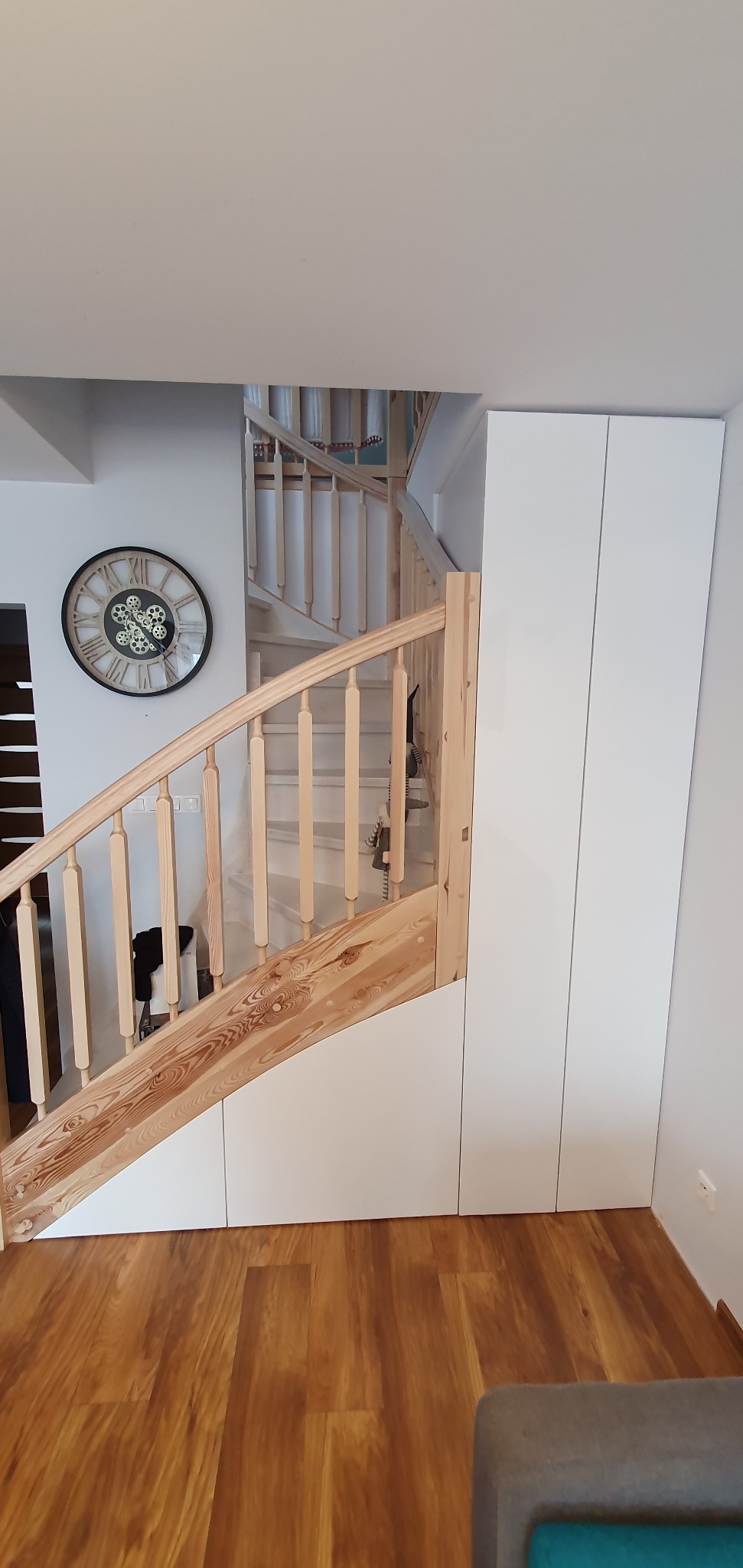 Drewniane schody spiralne z zabudową szaf wnękowych pod biegiem, obok ściany zegar w stylu industrialnym, na podłodze panele imitujące drewno.