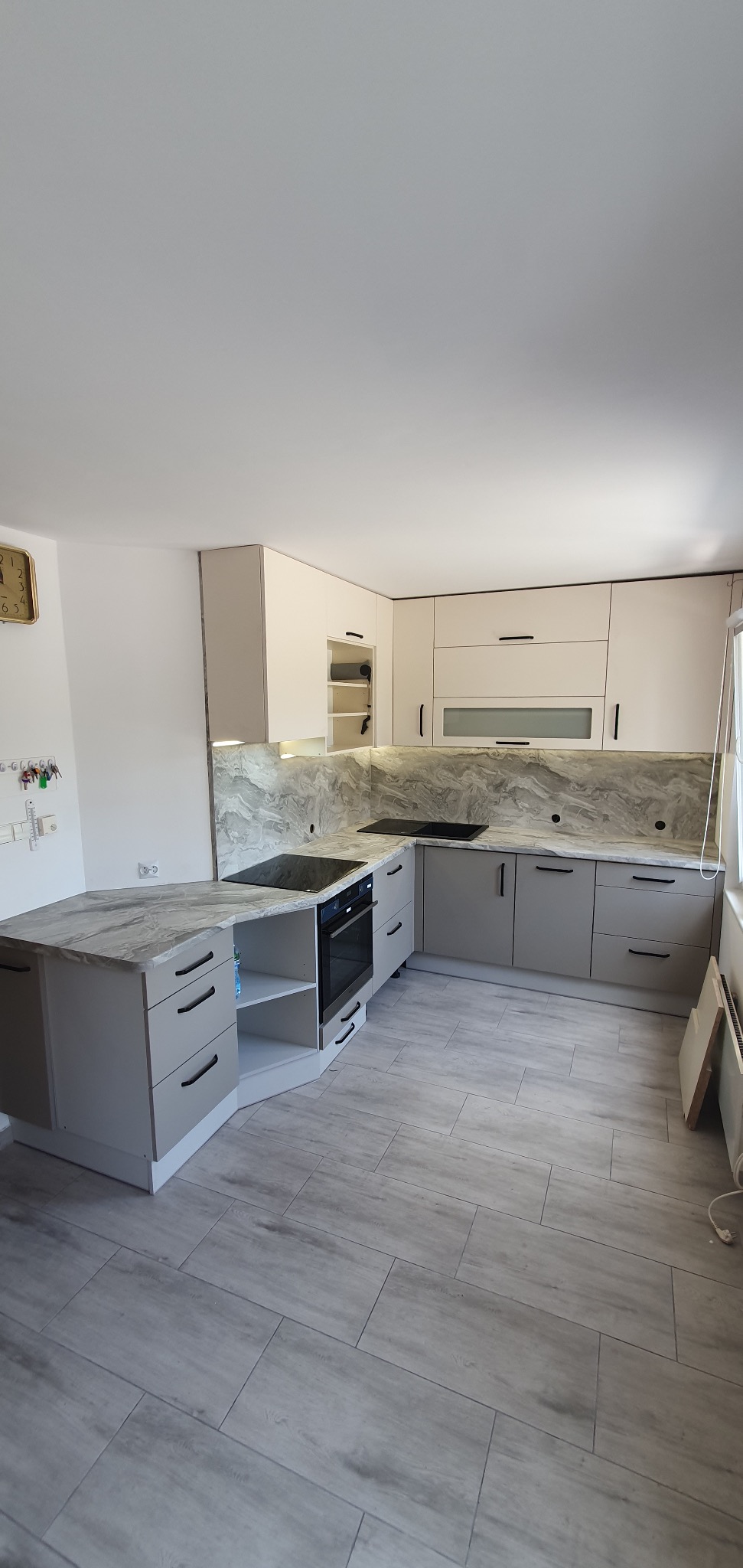 Jasna kuchnia z szarymi frontami i blatem imitującym marmur, czarne, podłużne uchwyty, płyta indukcyjna i piekarnik w zabudowie, widoczny zegar na ścianie.