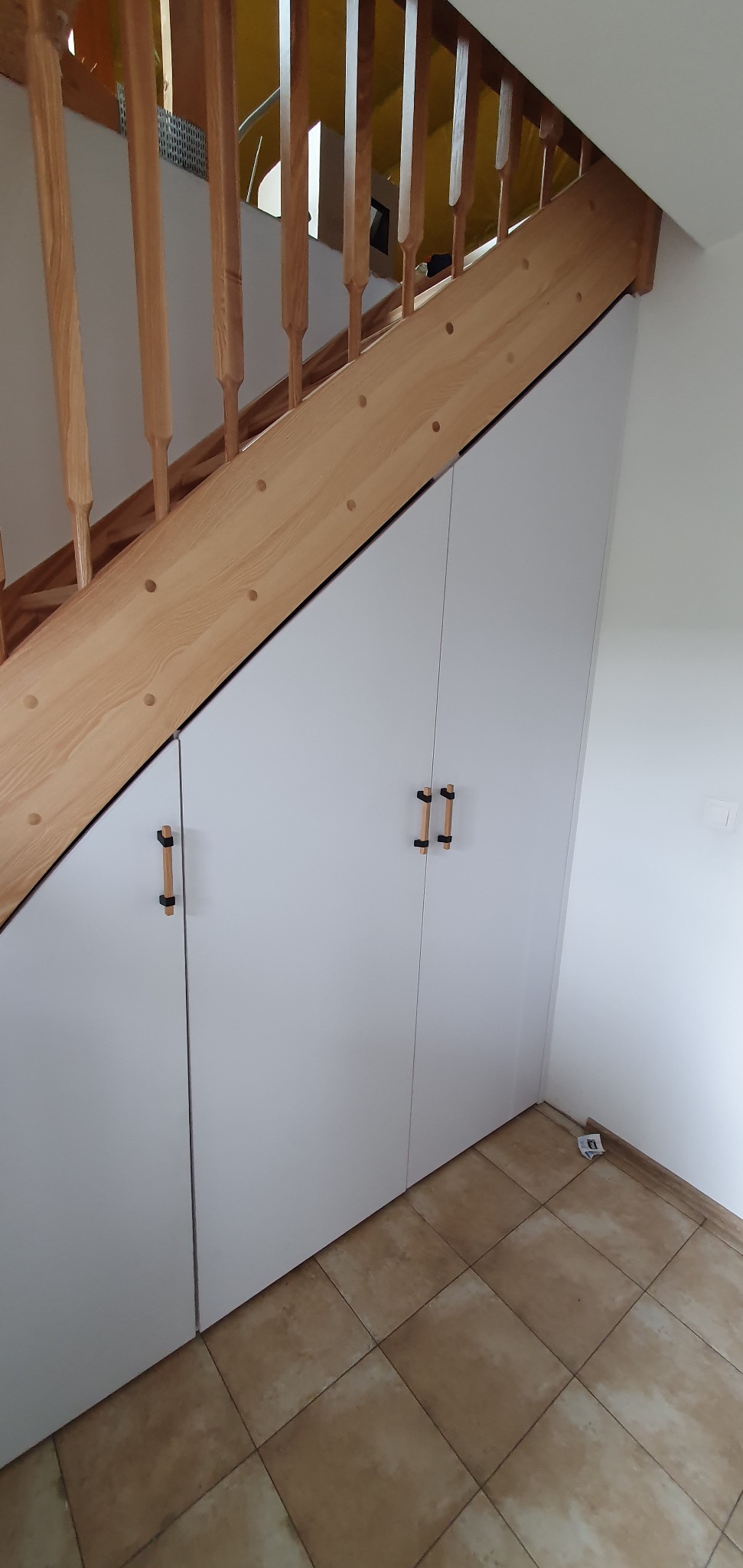 Biała szafa wnękowa pod schodami z drewnianymi uchwytami, zrealizowana w Tychach, dopasowana do skosu schodów z drewnianą balustradą.