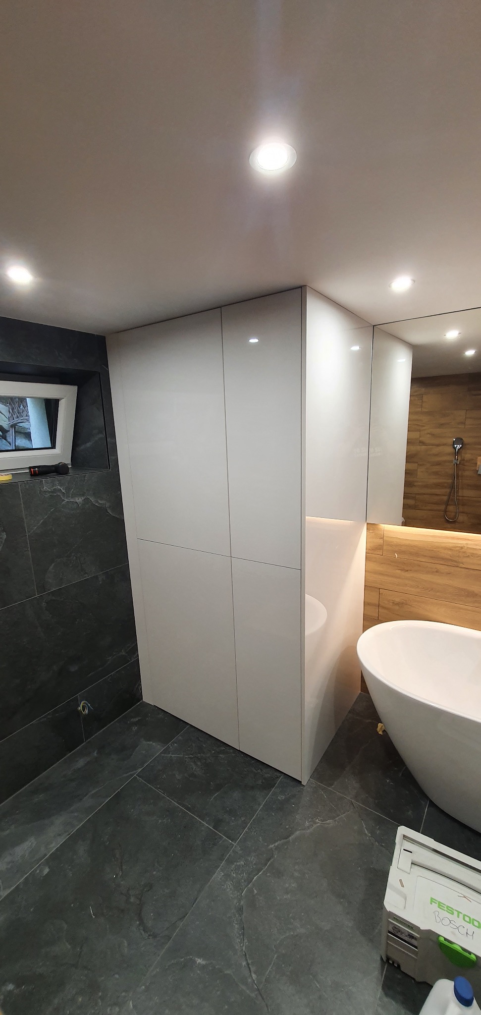 Biała szafa na wymiar w łazience z połyskiem, obok wolnostojąca wanna i drewnopodobna okładzina ściany, narzędzia na podłodze sugerują trwający montaż.