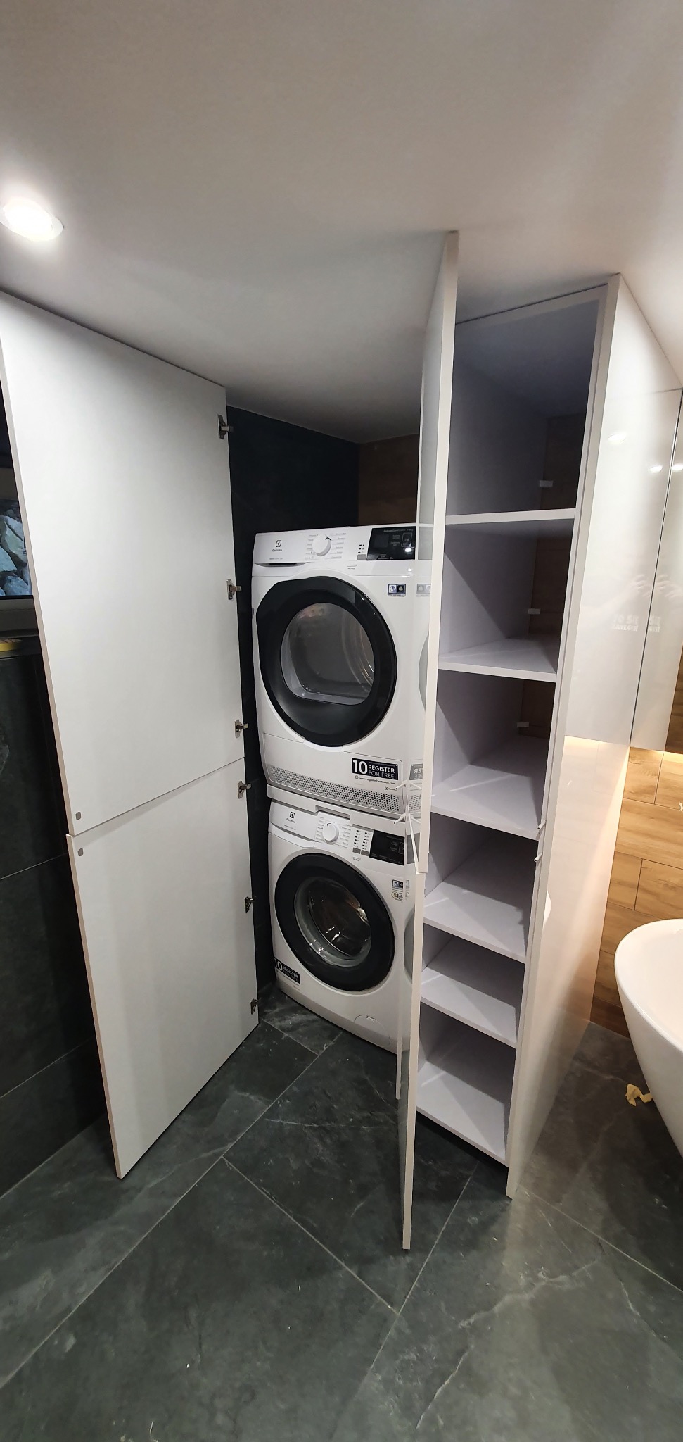 Biała zabudowa meblowa w łazience z pralką i suszarką w kolumnie, otwarte półki po prawej stronie i zamknięte szafki po lewej, szara podłoga z płytek.