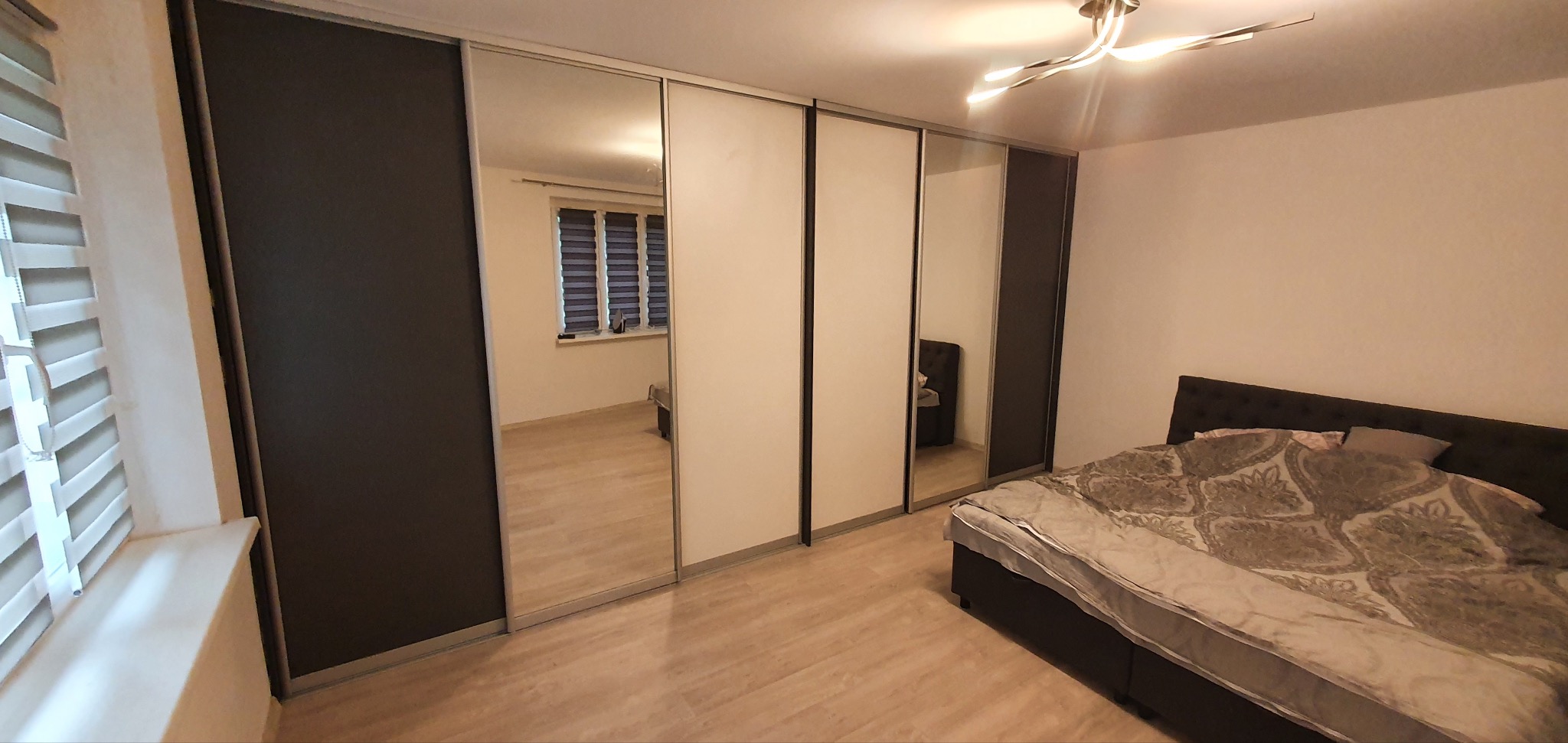 Nowoczesna szafa przesuwna z lustrami w sypialni, widoczne odbicie okna z roletami i fragment łóżka z wzorzystą pościelą.