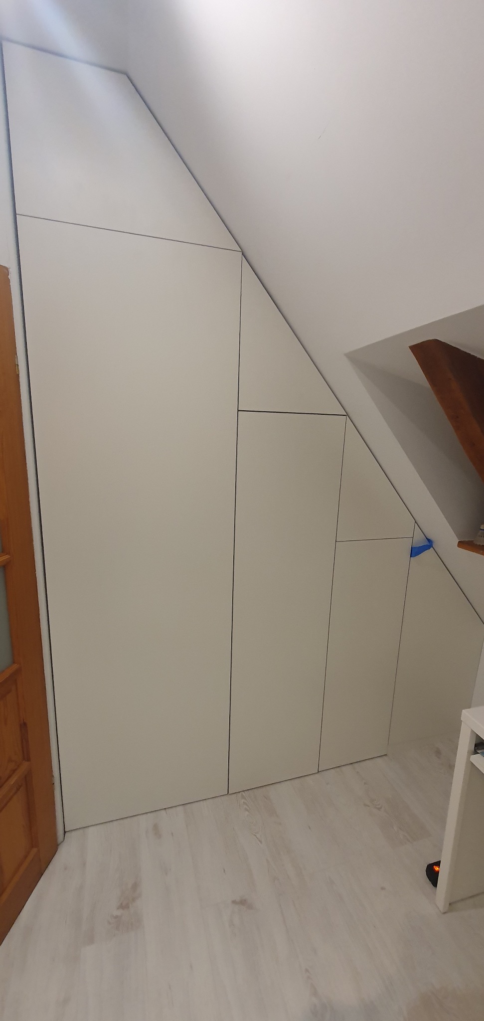 Biała szafa wnękowa na poddaszu, dopasowana do skosu dachu, widoczne drzwi i fragment drewnianych drzwi po lewej stronie.