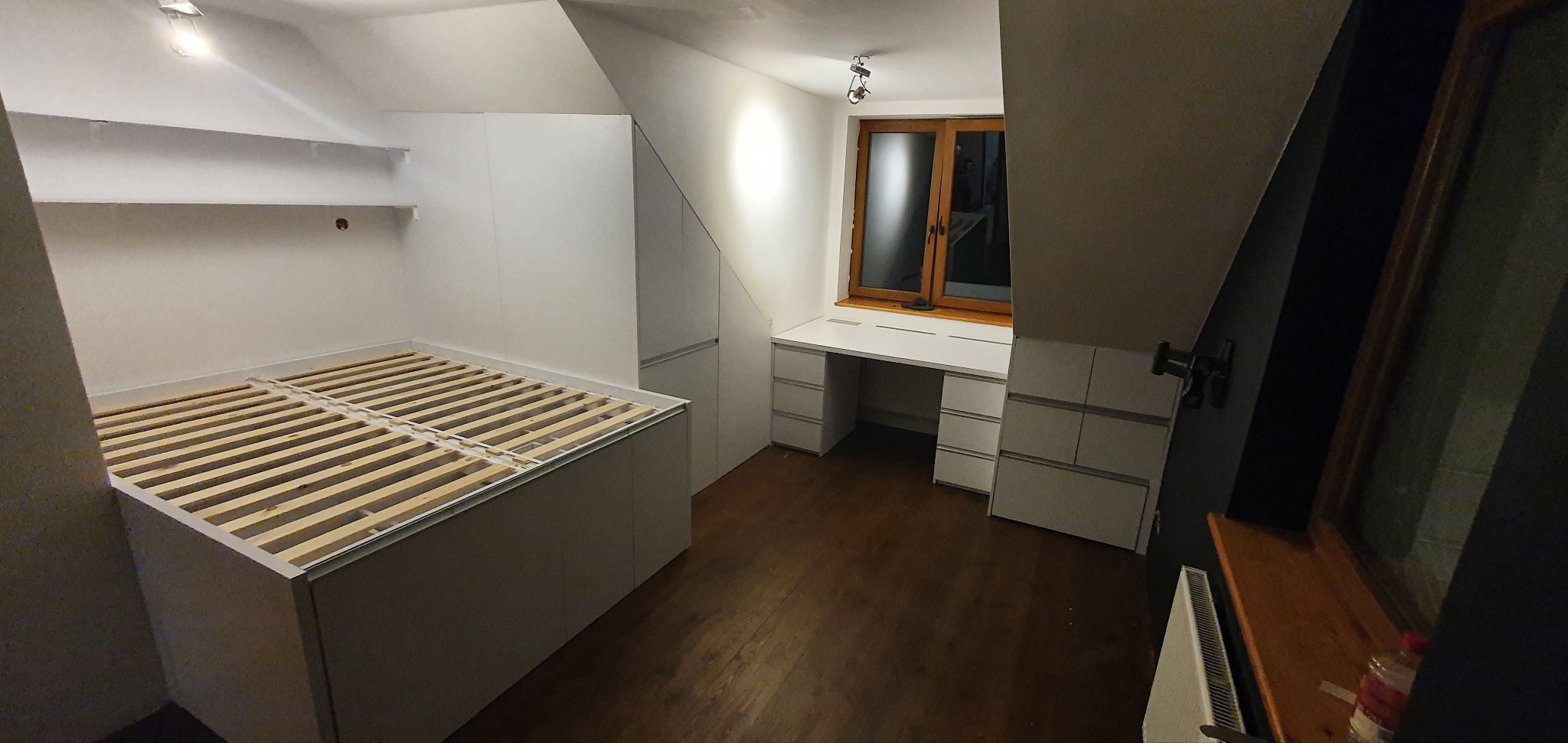 Pokój na poddaszu z łóżkiem na podeście z drewnianym stelażem, białą zabudową szaf i biurkiem przy oknie.