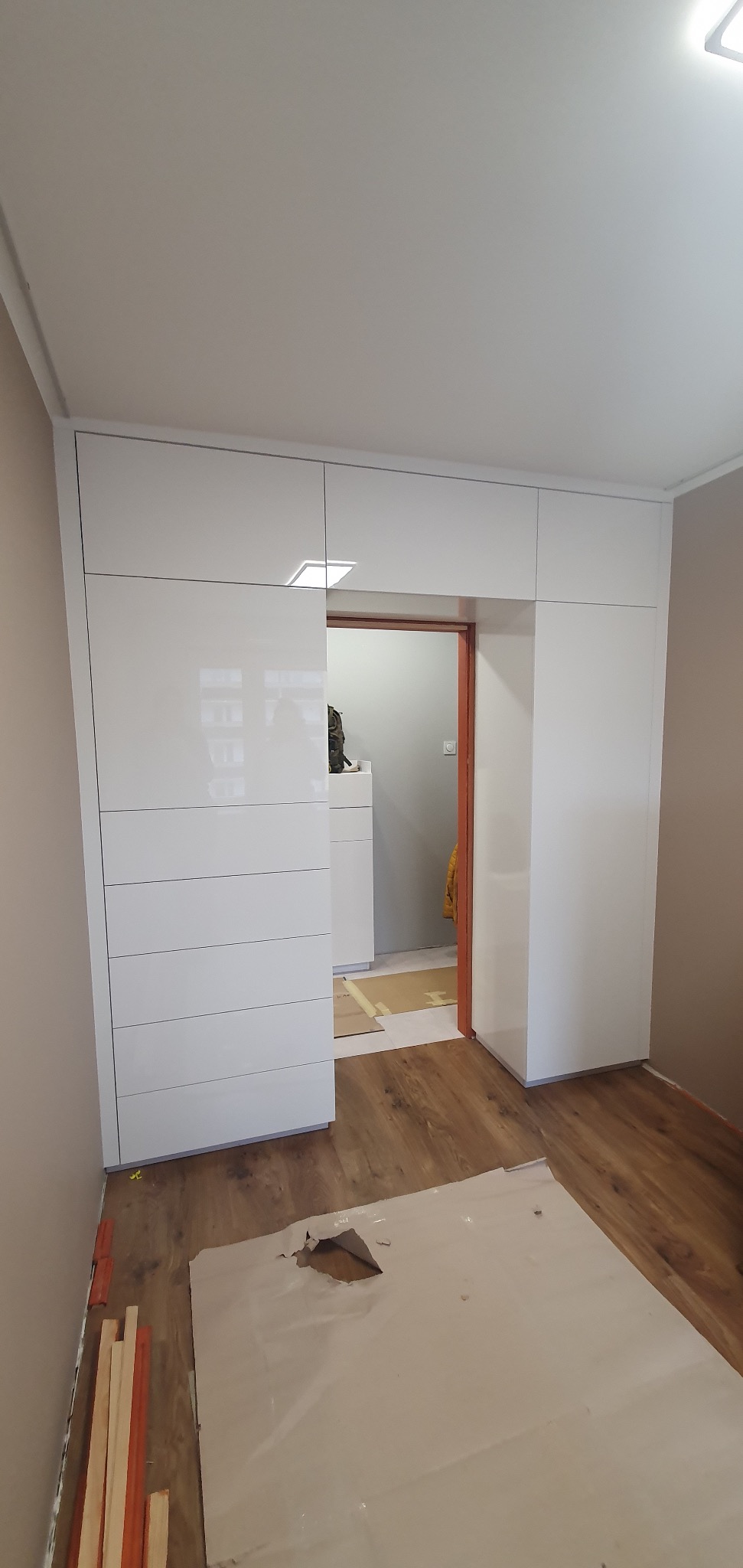 Biała, błyszcząca szafa wnękowa z przesuwnymi drzwiami, widok na wnętrze garderoby przez otwarte drzwi, podłoga zabezpieczona kartonem, listwy przypodłogowe przy ścianie.