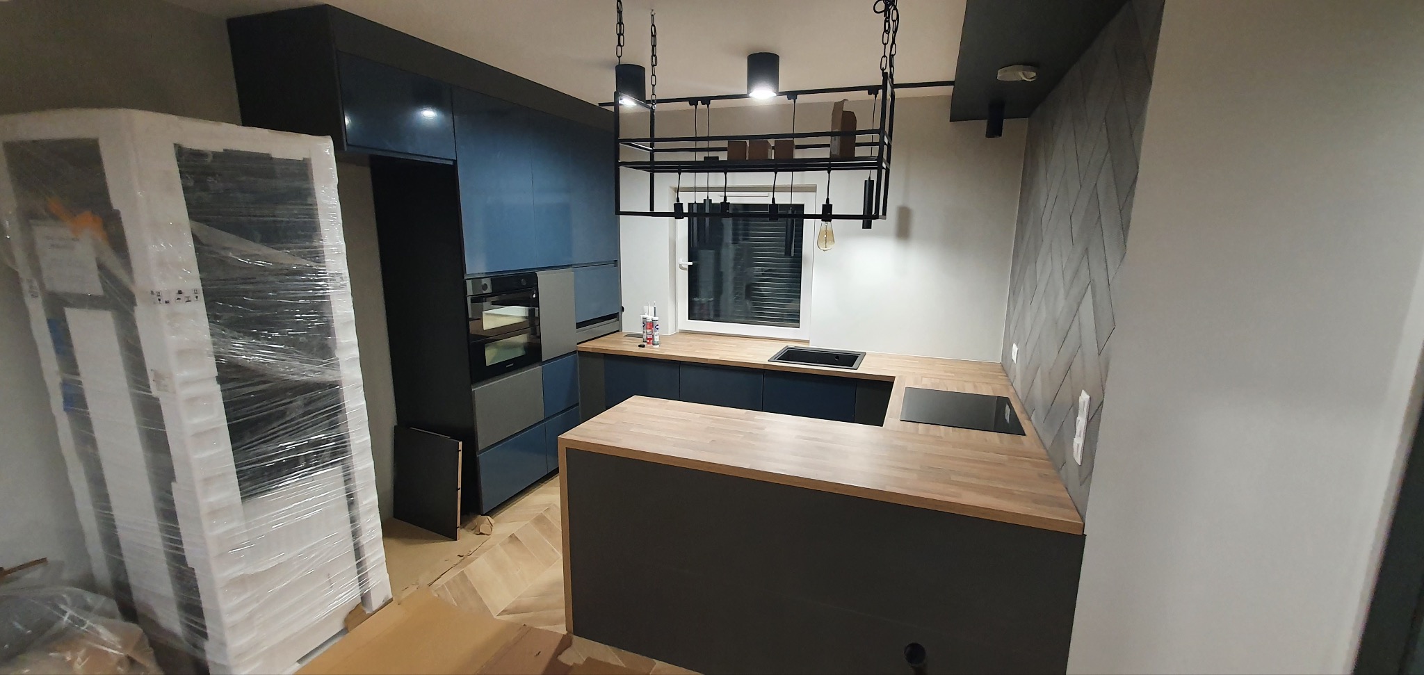 Niebiesko-szara zabudowa kuchenna z drewnianym blatem, wyspą i czarnym oświetleniem wiszącym, częściowo jeszcze w folii ochronnej.