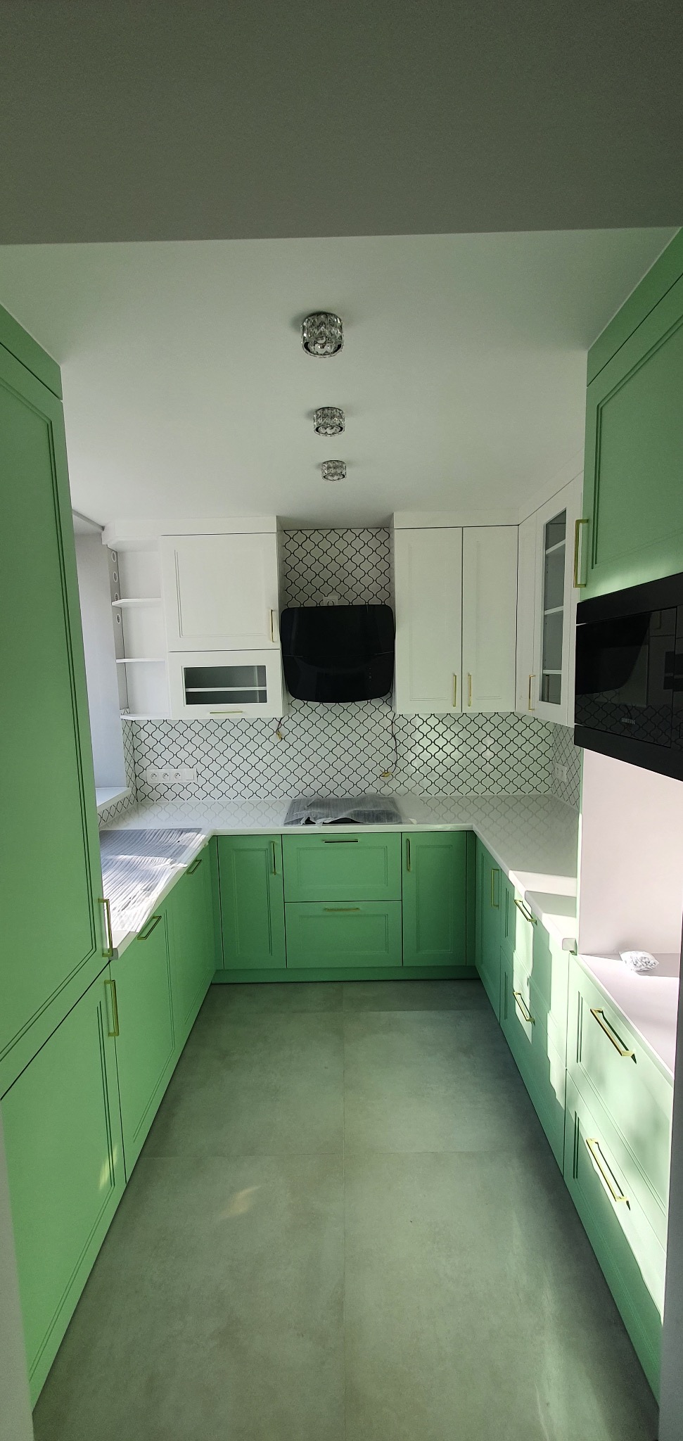 U-kształtna kuchnia z zielonymi szafkami dolnymi i białymi górnymi, złote uchwyty, czarny okap, płytki w marokański wzór, trzy kryształowe lampy sufitowe.