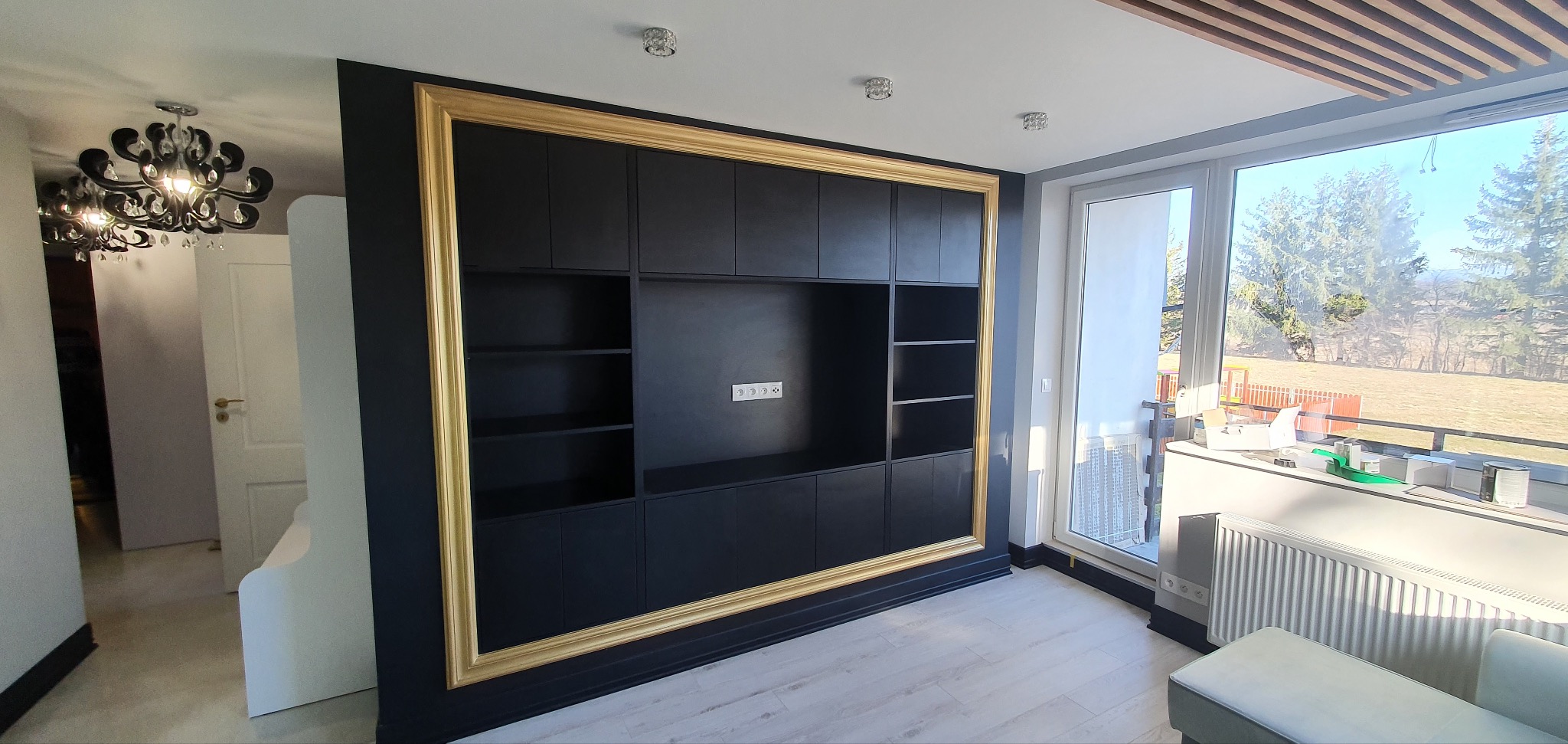 Czarna zabudowa meblowa z półkami i szafkami, obramowana złotą ramą, w nowoczesnym wnętrzu z widokiem na zewnątrz.