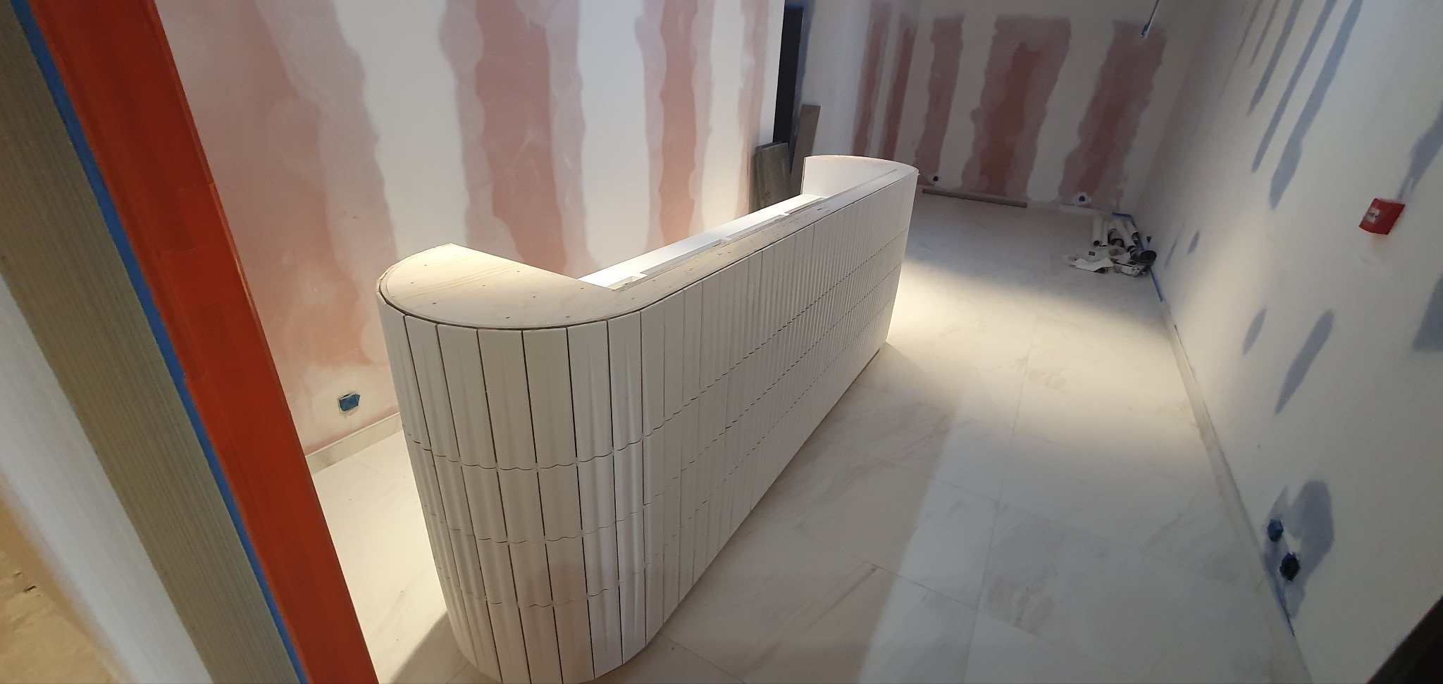 Biała, zaokrąglona lada recepcyjna z pionowymi żłobieniami, w surowym stanie, w trakcie montażu w pomieszczeniu z przygotowanymi ścianami do malowania.