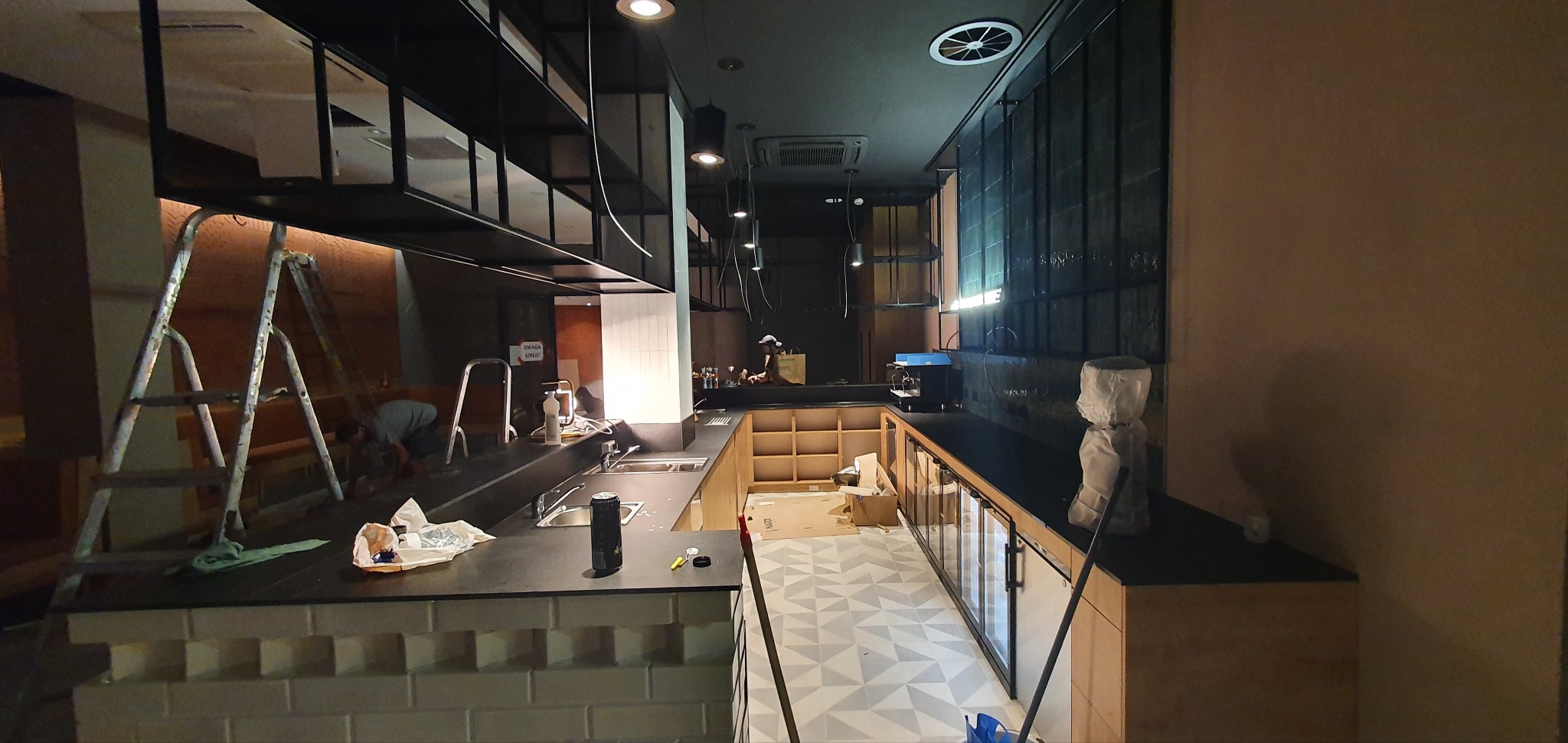 Montaż nowoczesnej kuchni w lokalu gastronomicznym: widoczne niedokończone szafki, blaty robocze, instalacja oświetlenia, pracownik na drabinie i częściowo zakryta podłoga z wzorem geometrycznym.