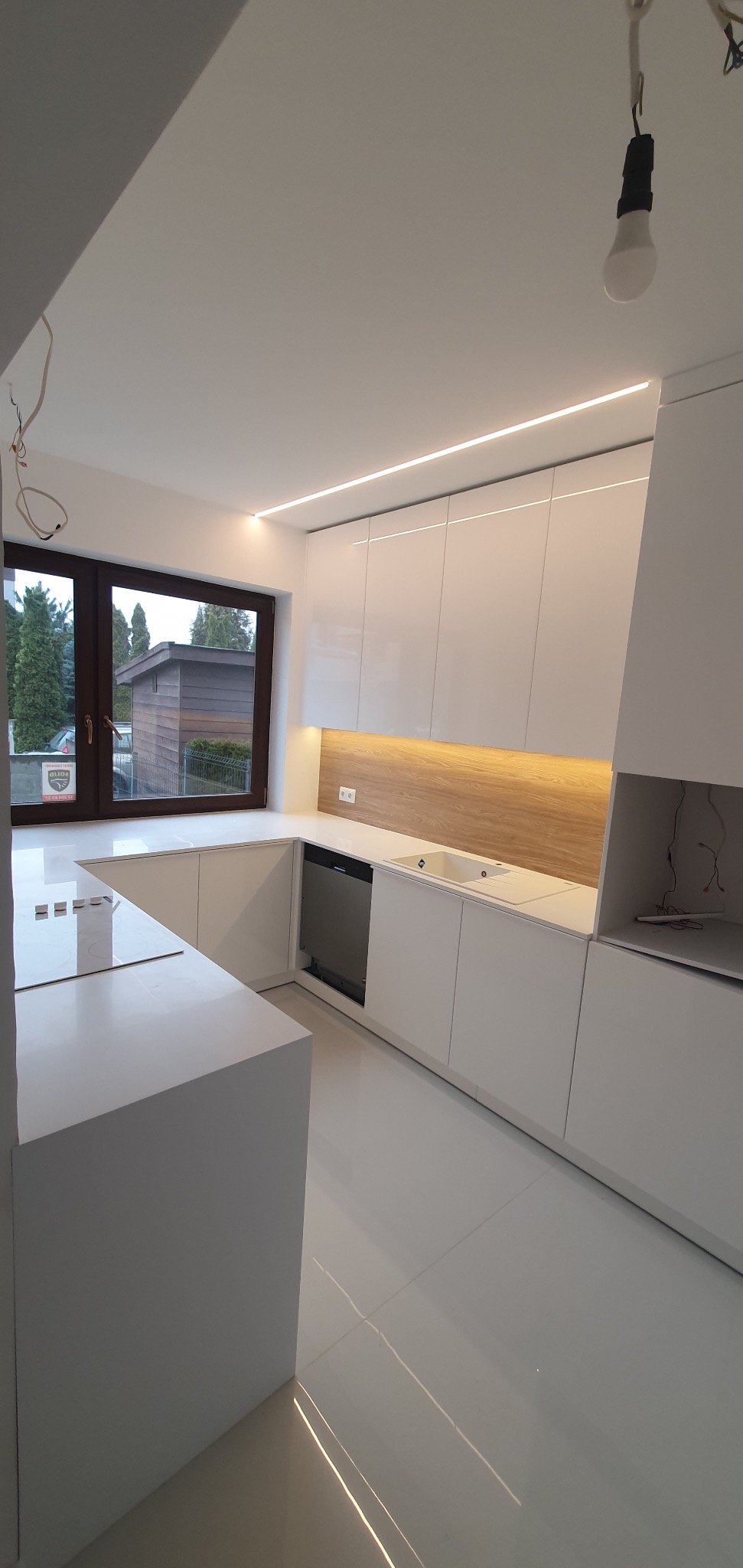 Nowoczesna, biała kuchnia z drewnianym panelem nad blatem, oświetleniem LED podszafkowym i dużym oknem z widokiem na zieleń, z niedokończonymi pracami elektrycznymi widocznymi na suficie.