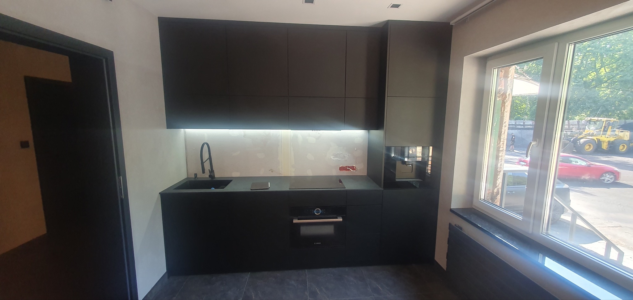 Ciemna, minimalistyczna zabudowa kuchenna z czarnym zlewem, płytą indukcyjną i piekarnikiem Bosch. Ściana nad blatem roboczym w trakcie wykończenia. Za oknem widoczny sprzęt budowlany i czerwony...