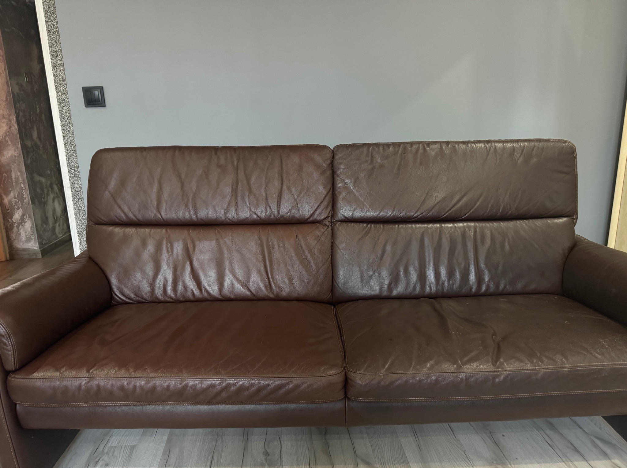 Dwuosobowa, brązowa, skórzana kanapa z widocznymi śladami użytkowania na siedzisku, ustawiona na panelach podłogowych, na tle szarej ściany z włącznikiem światła.