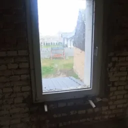 Surowa ceglana ściana z oknem PCV w trakcie montażu, widoczne drewniane podpory i zarysowany parapet, na zewnątrz widok na podw&oacute;rko z huśtawką.