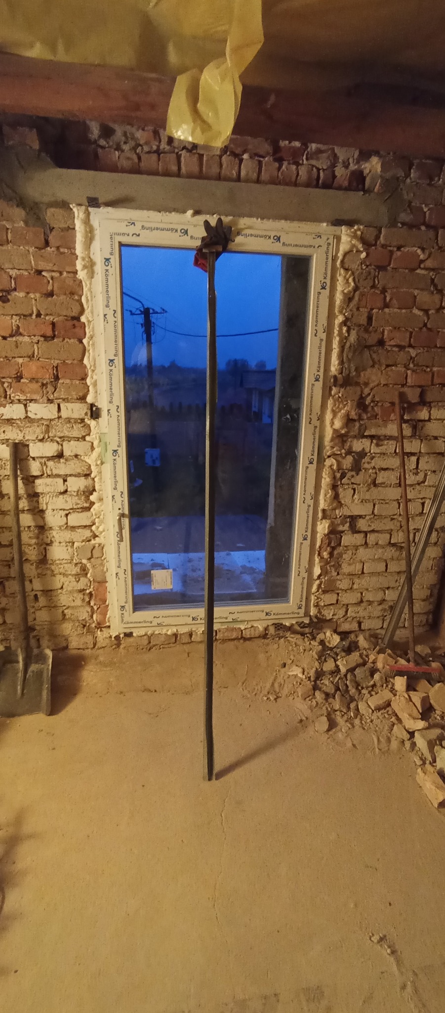 Montaż okna PCV w surowym murze z cegły, uszczelnionego pianką montażową, z widocznym narzędziem do podtrzymywania ramy i widokiem na zewnątrz o zmierzchu.