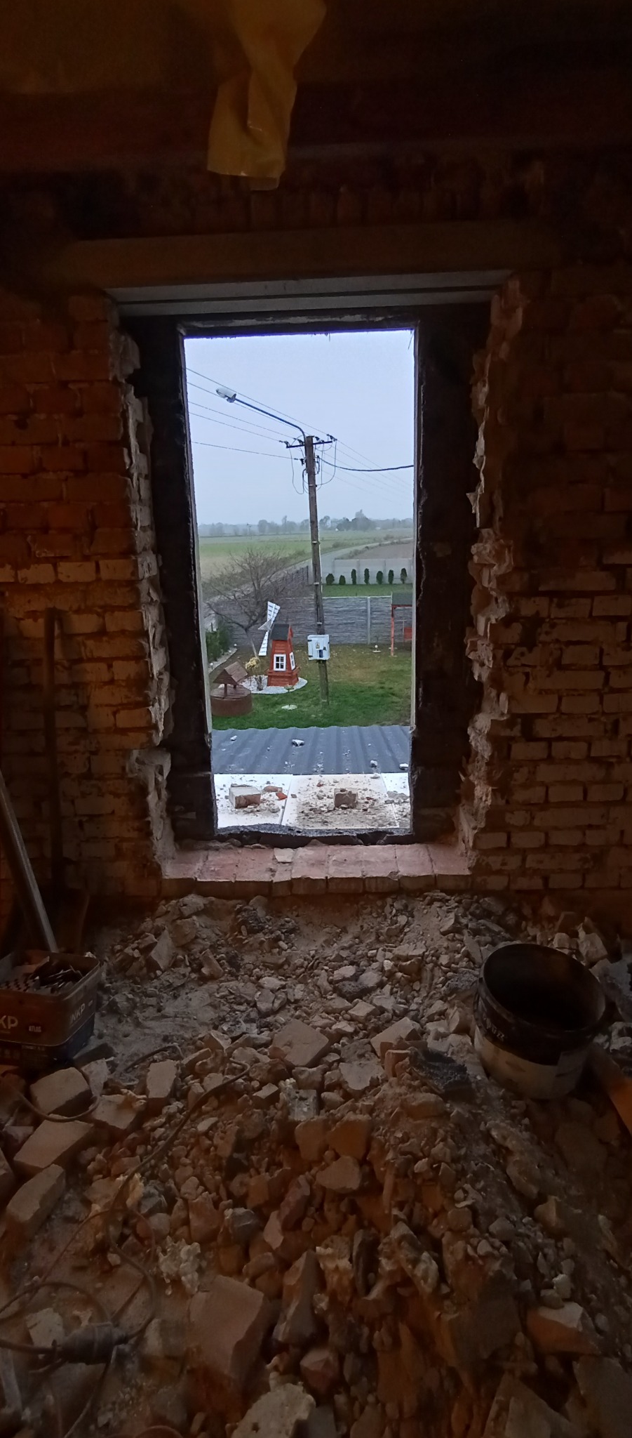 Otwór okienny w trakcie wykuwania w ścianie z czerwonej cegły, widok na zewnątrz z małym wiatrakiem ogrodowym i słupem elektrycznym