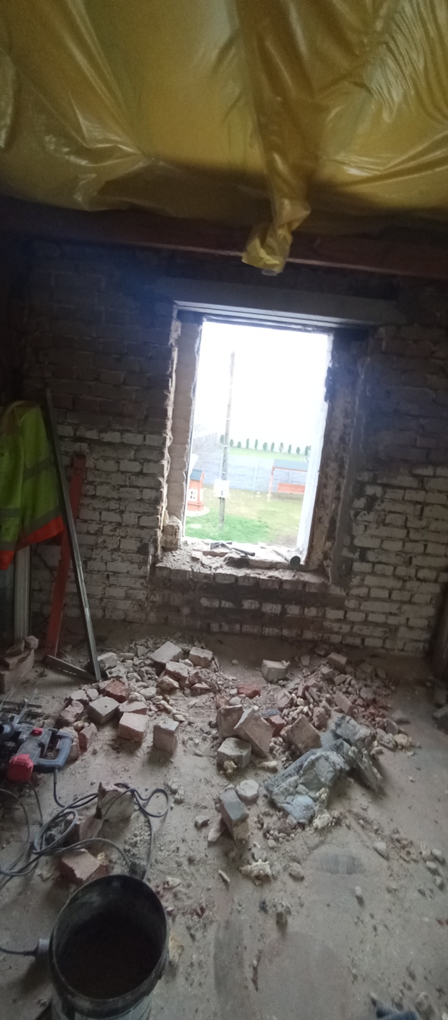 Wnętrze w trakcie demontażu muru okiennego z cegły, widoczne gruz, narzędzia i zabezpieczenie folią. Za oknem zarys krajobrazu.
