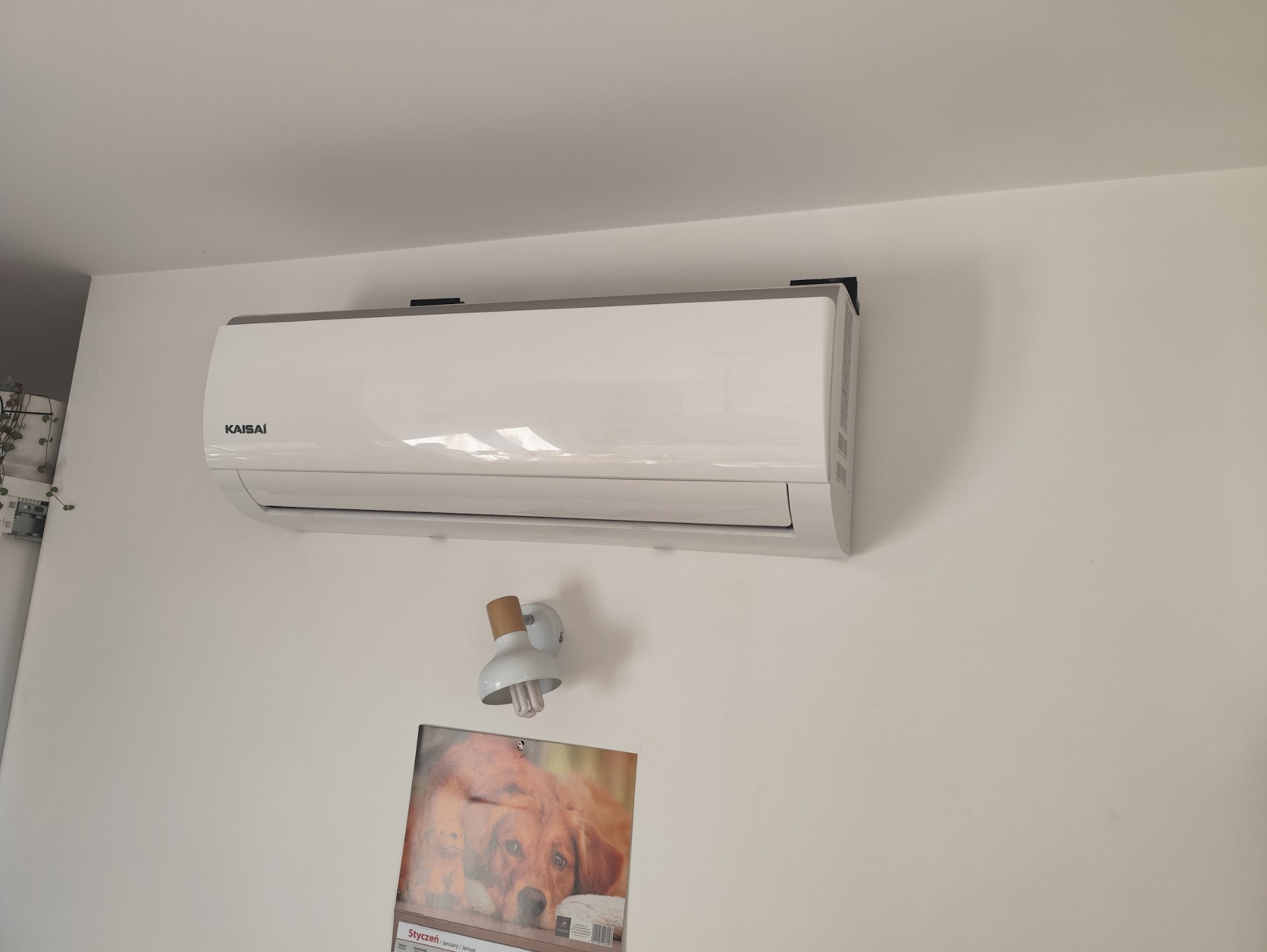 Biały klimatyzator Kaisai zamontowany na ścianie, widoczna lampka i kalendarz. Minimalistyczny design, czyste linie, neutralne tło.