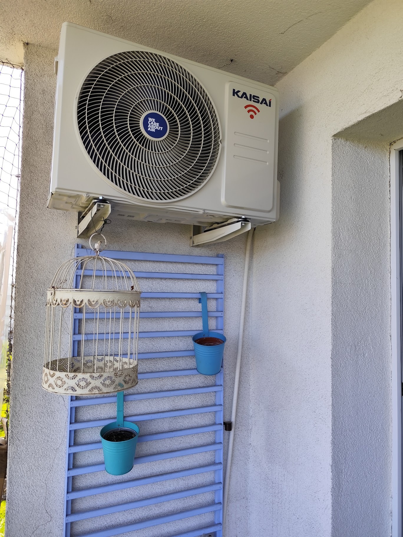 Biały klimatyzator Kaisai zamontowany na balkonie z dekoracyjną kratką i doniczkami. Widoczne detale montażu i estetyczne wkomponowanie w przestrzeń.